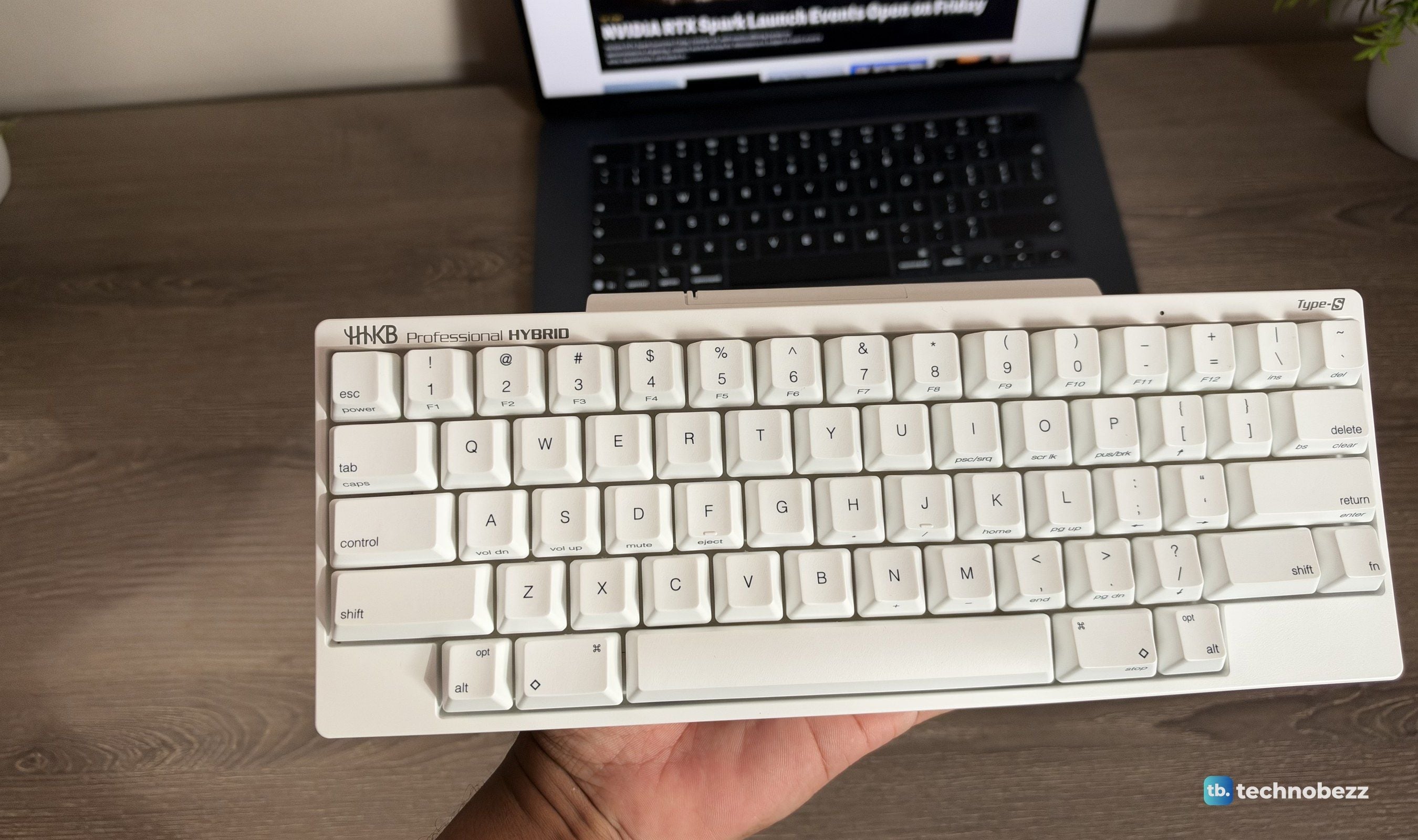 HHKB held in one hand in front of a MacBook on a wooden desk