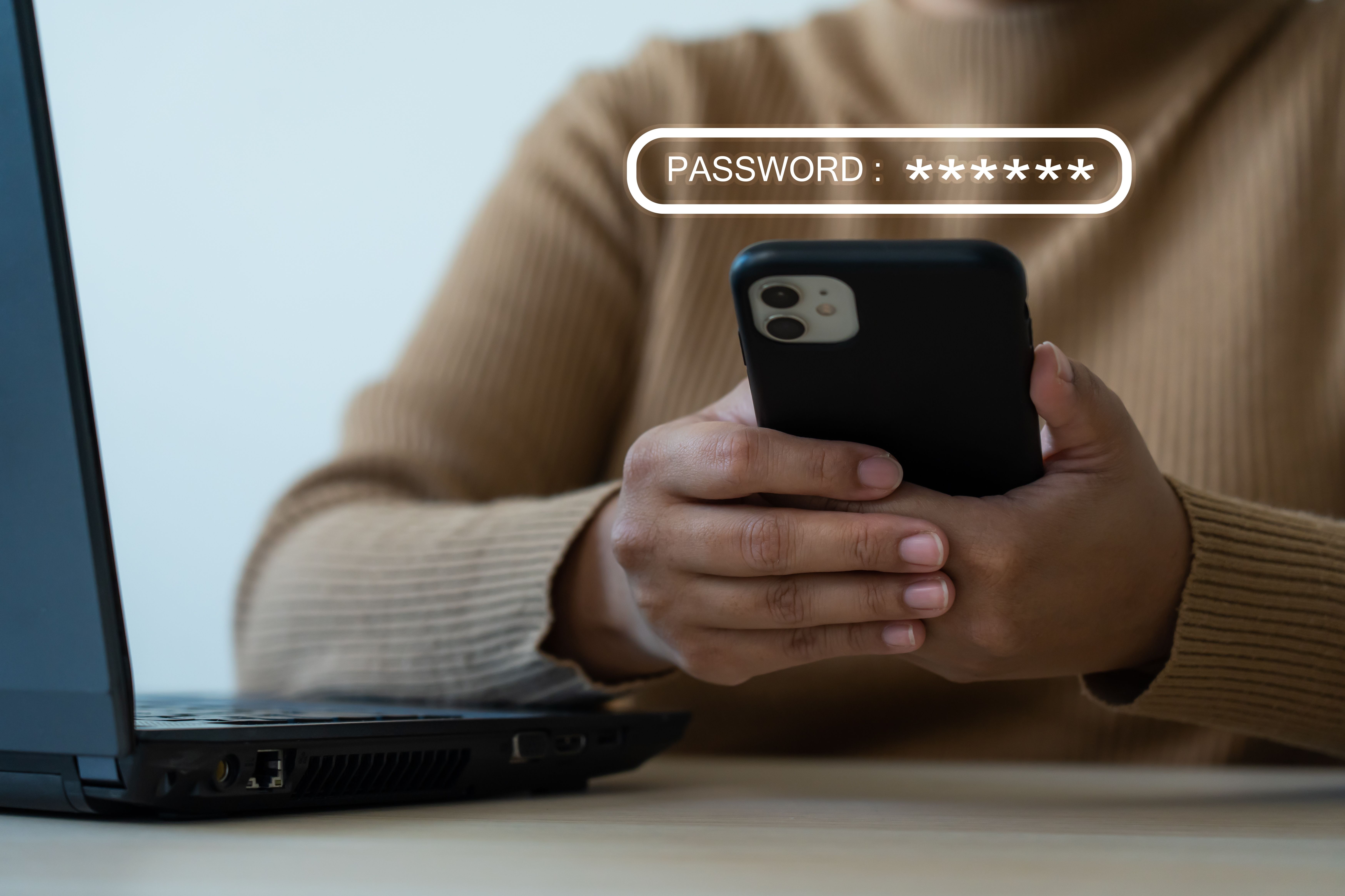 Hands holding a smartphone in front of a laptop with a password field above it