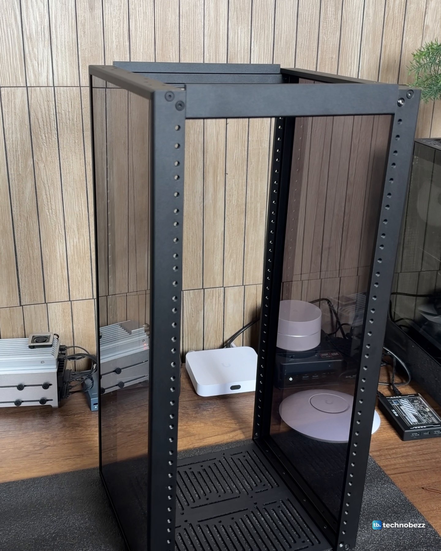 An empty black 12U rack frame with smoked acrylic side panels standing on a wood desk, with a white gateway and small boxes behind it