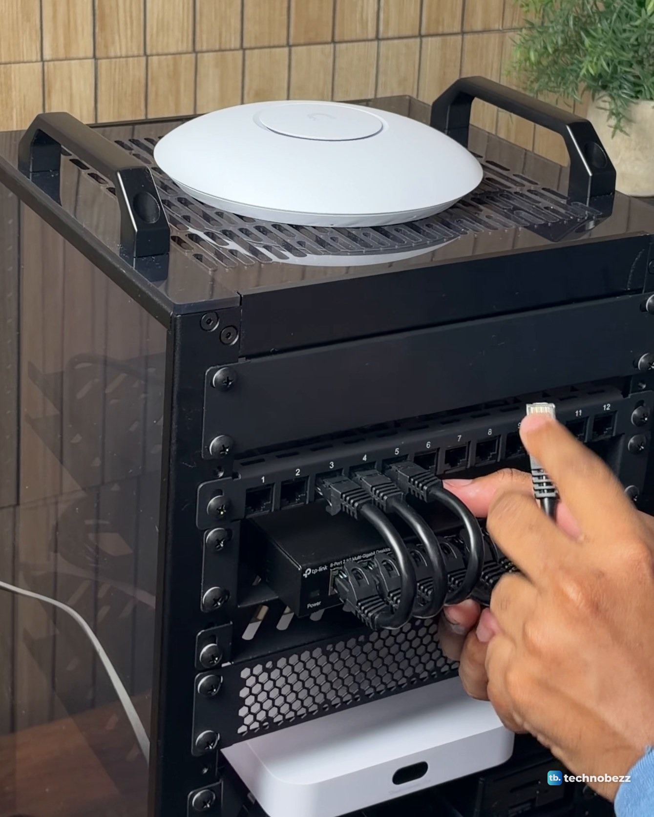 A hand plugging a short black Ethernet jumper into the 12-port patch panel above the TP-Link switch, with the access point on top of the rack
