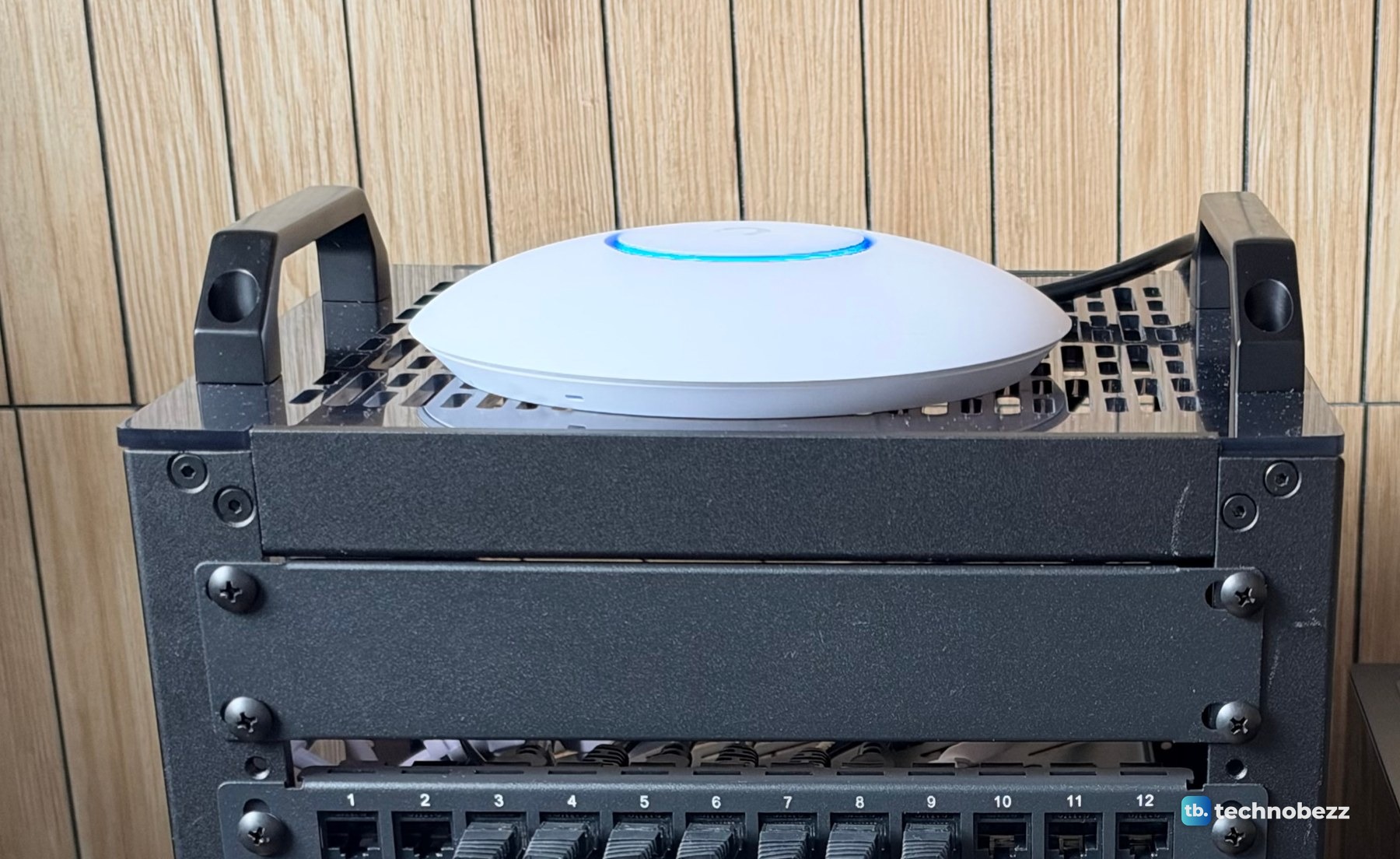The round white UniFi U7 Lite access point lying on the vented top plate of the rack between two black handles