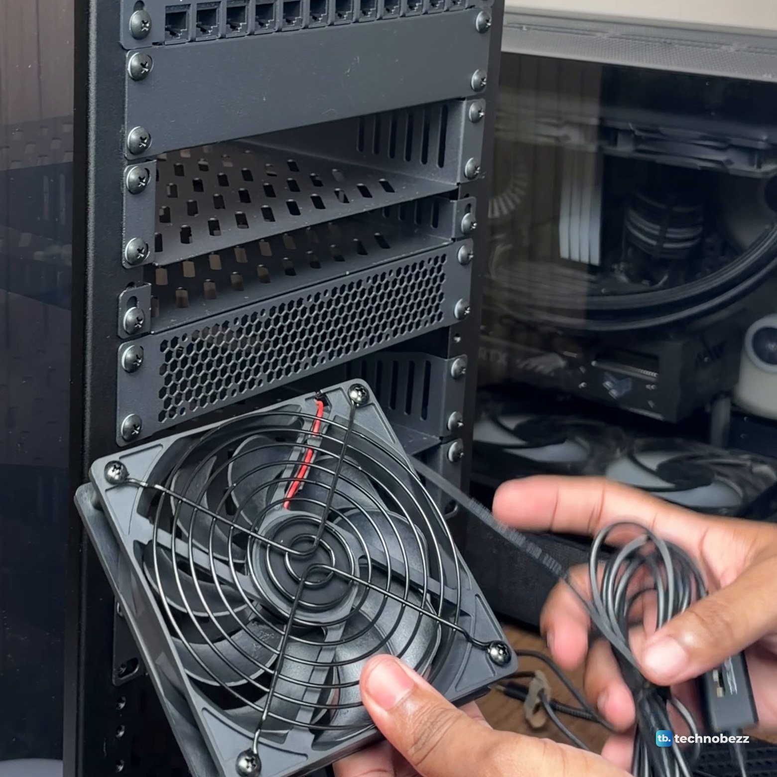 A hand holding a black 120 mm fan with a wire grille and a USB cable in front of the rack
