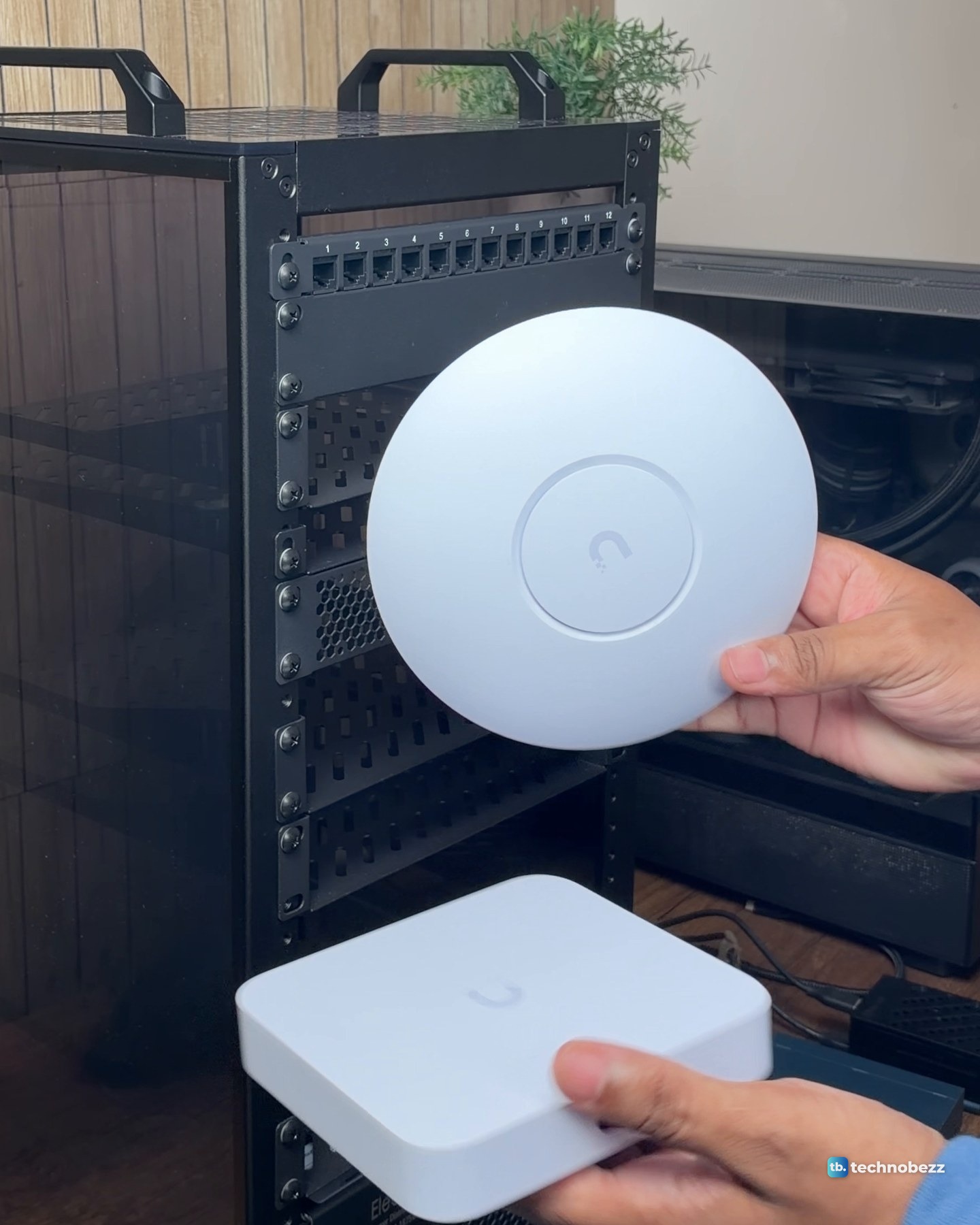 Hands holding the round white UniFi U7 Lite above the white UniFi Cloud Gateway Ultra in front of the rack