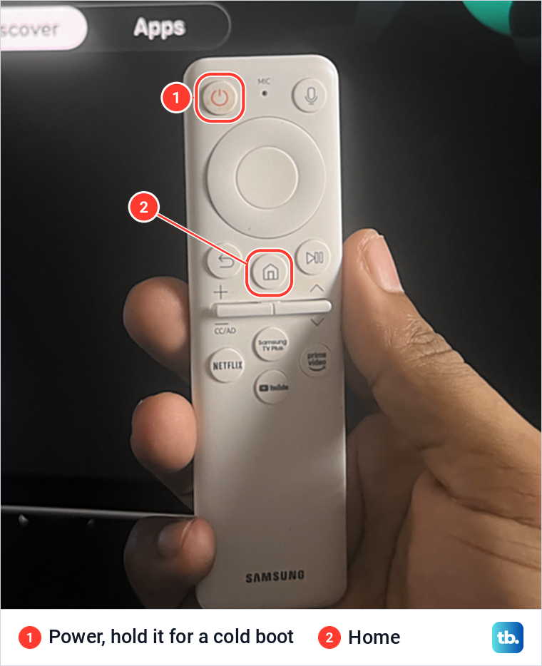 A hand holding a white Samsung remote with the Power button at the top left and the Home button marked.
