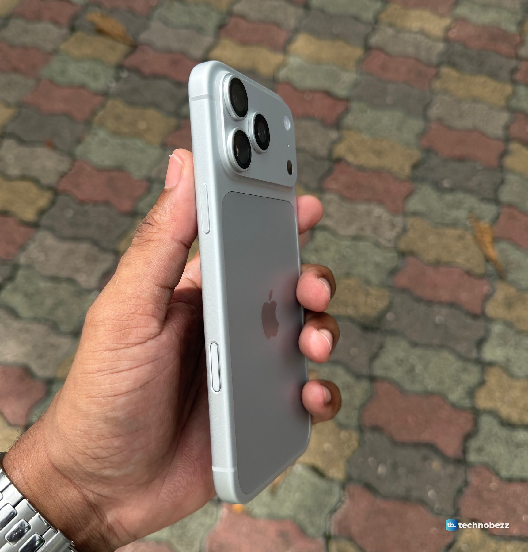 Back and right edge of a silver iPhone 18 Pro Max held in a hand, showing a raised button near the top of the edge and a flush control lower down