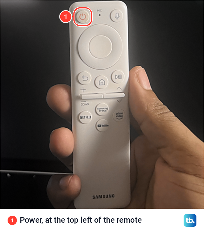 A hand holding the white Samsung remote with the power button at its top left marked.