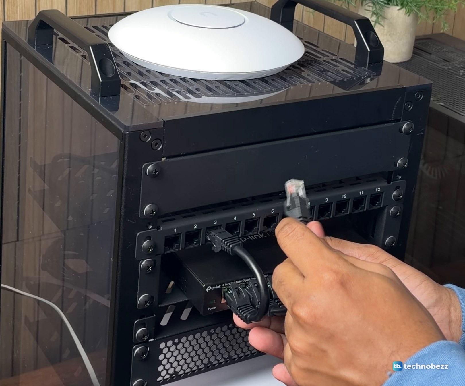 Hand holding a short black Ethernet patch cable up to a patch panel above a switch