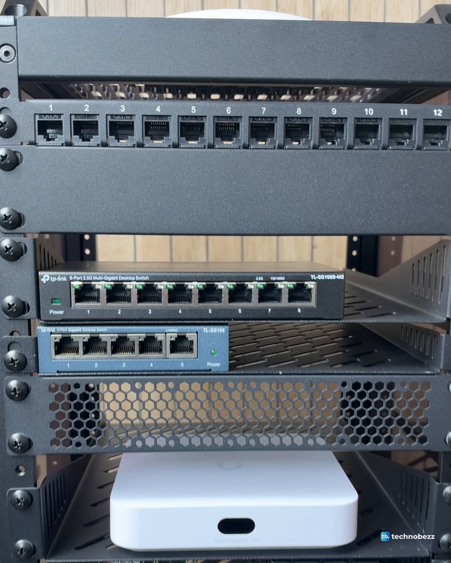 Front of the rack during loading: blank panels above and below the numbered 12-port patch panel, TP-Link 8-port and 5-port switches, a vented panel and the white UniFi Cloud Gateway Ultra on a shelf