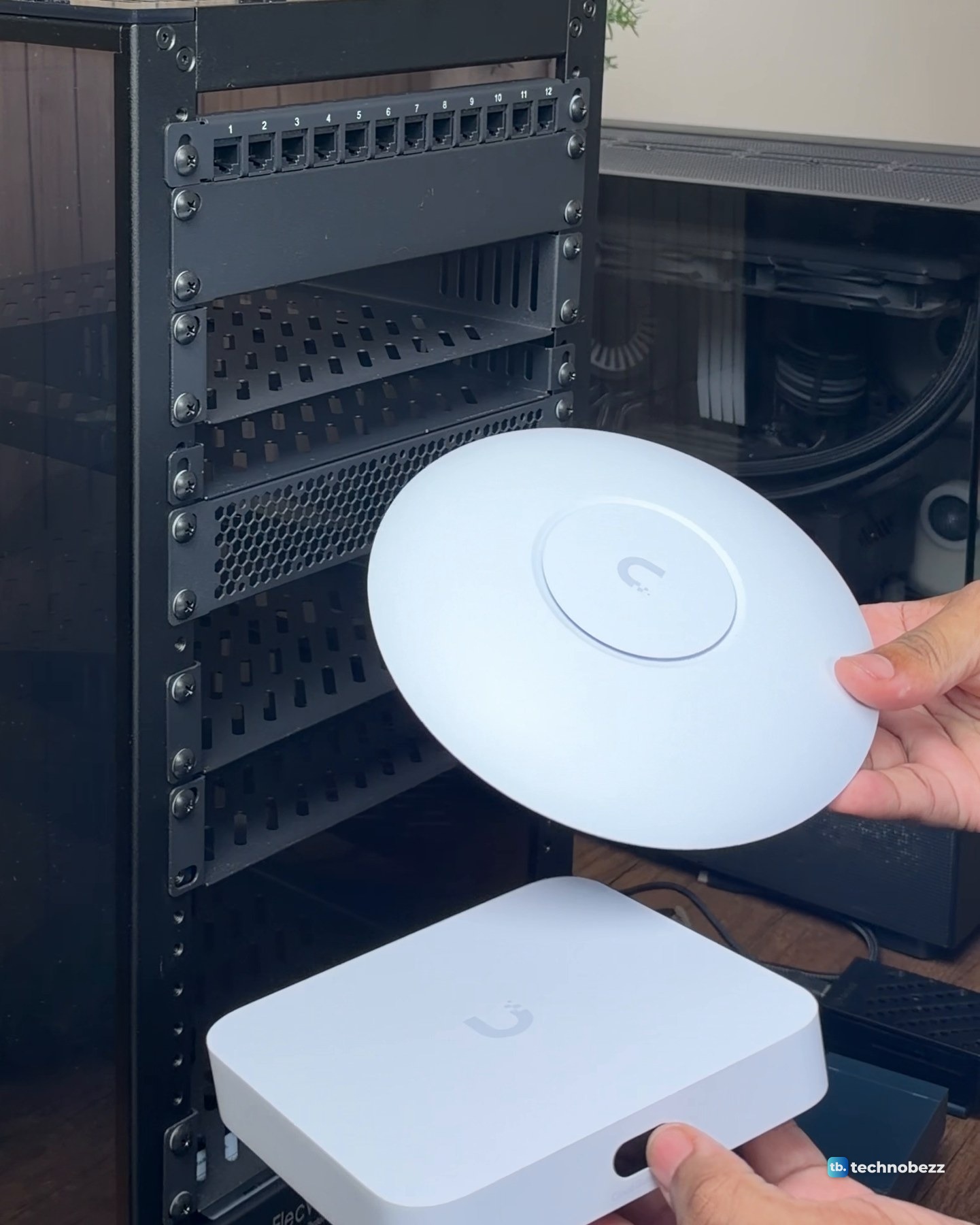 A hand holding the round white UniFi U7 Lite above the square white UniFi Cloud Gateway Ultra in front of the empty rack