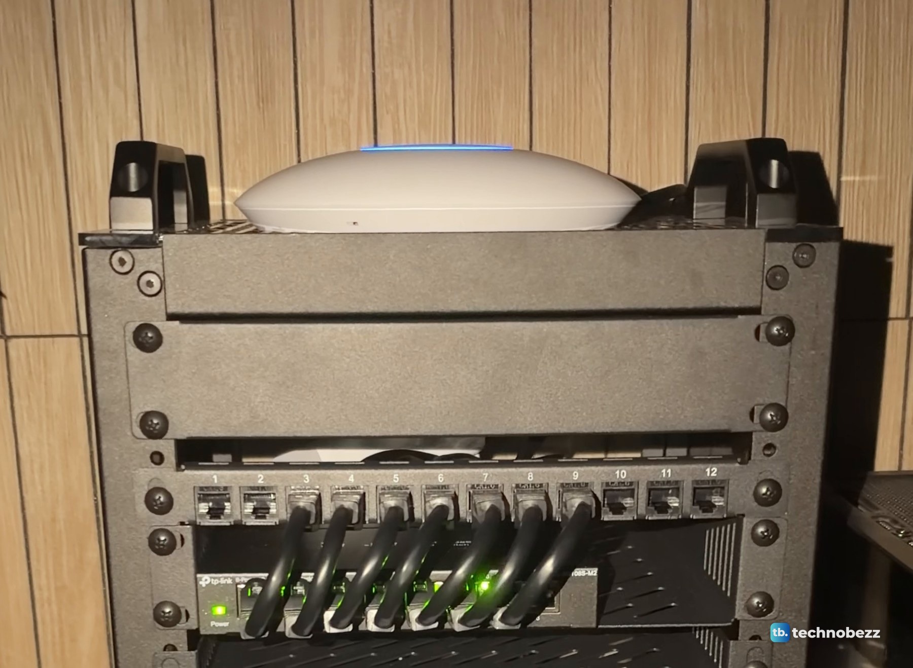 The rack in dim warm light with the U7 Lite on top glowing a thin blue ring and green link lights on the switch below the patch panel