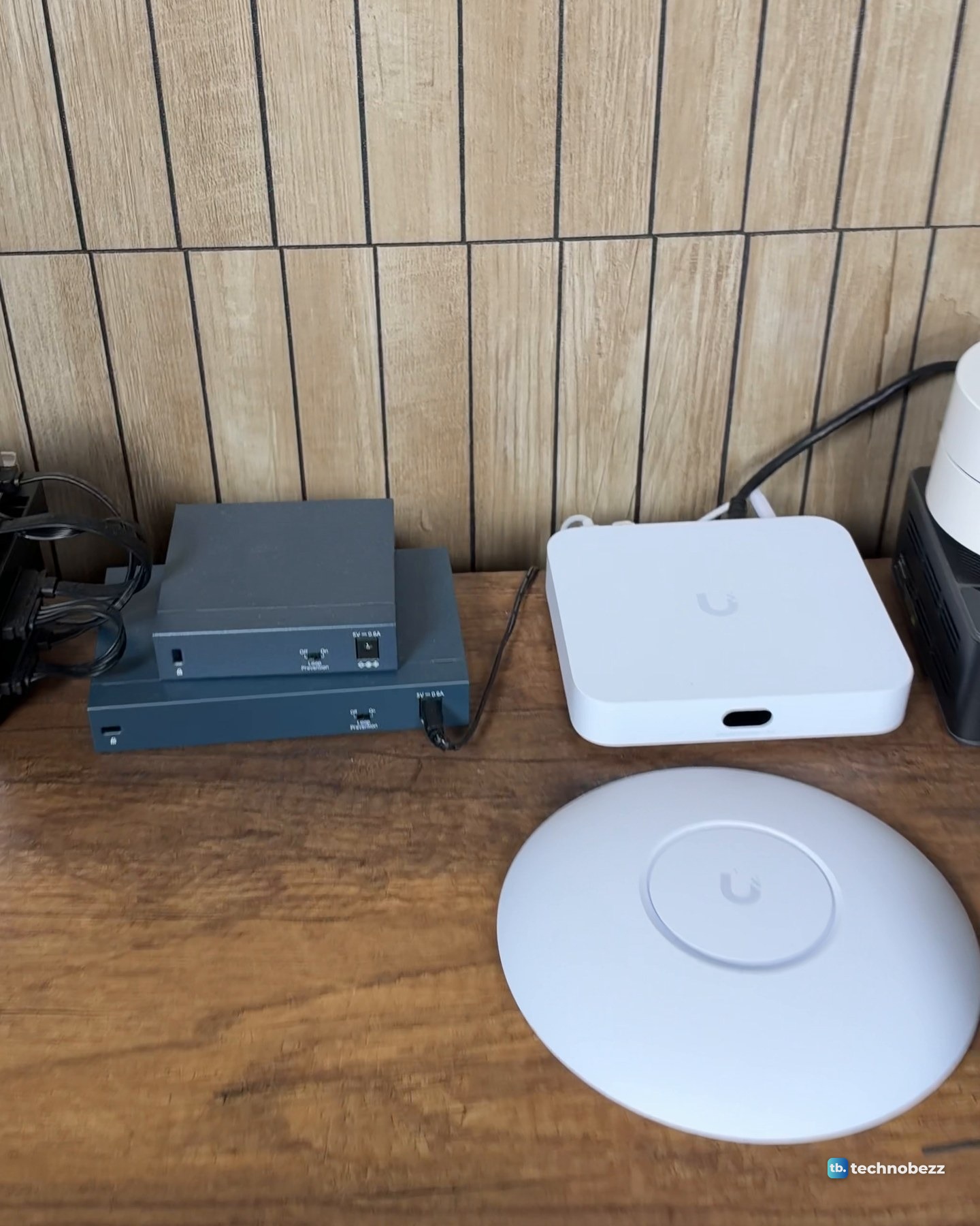UniFi U7 Lite access point lying flat on a wood desk beside a white UniFi Cloud Gateway Ultra and blue-grey network boxes