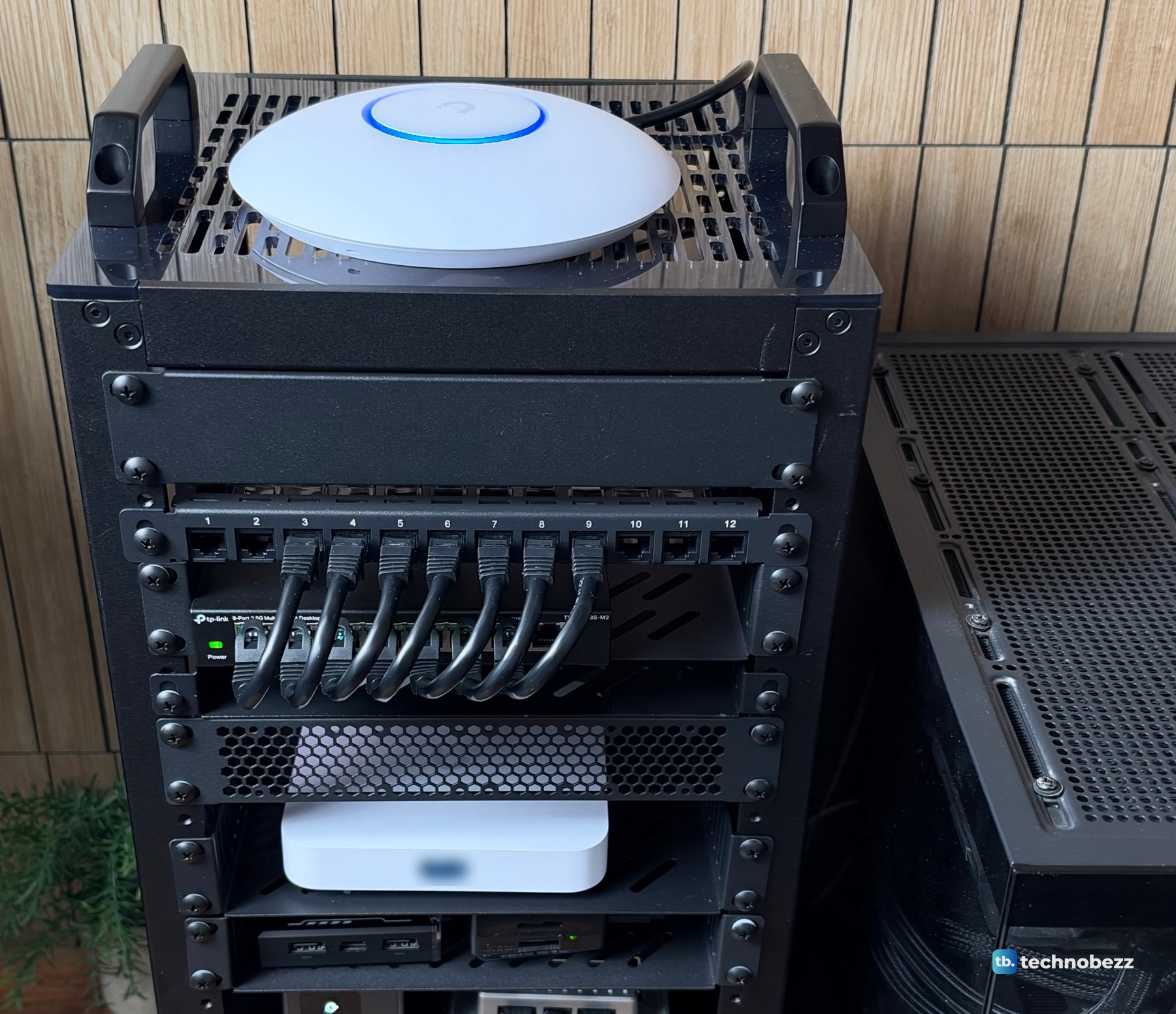 UniFi U7 Lite sitting on the vented top plate of the black rack, with a thin blue light ring around its logo and seven black patch cables in the panel below