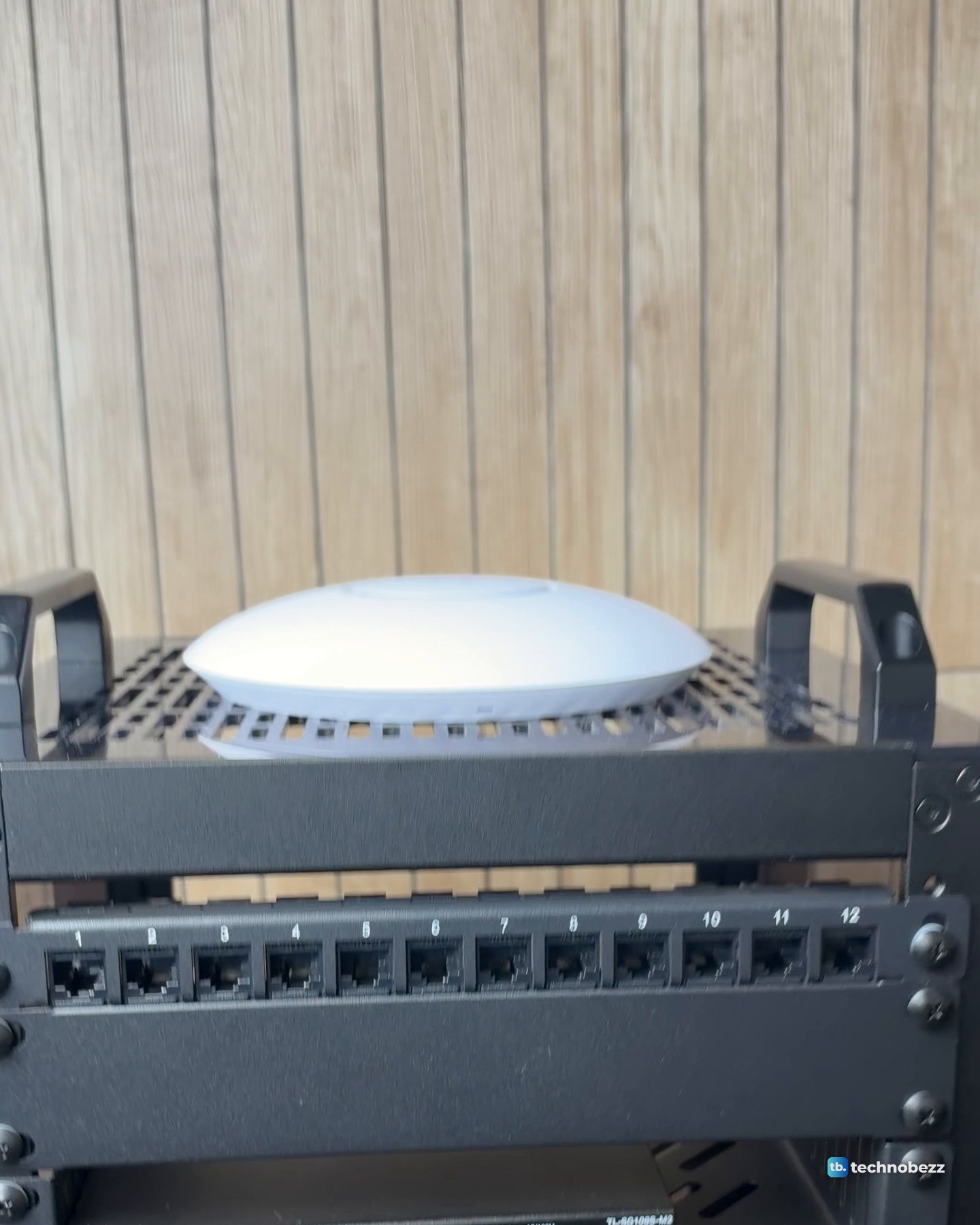 Close view of the white U7 Lite resting on the vented top plate between the rack's two black handles, above a blank panel and the numbered patch panel