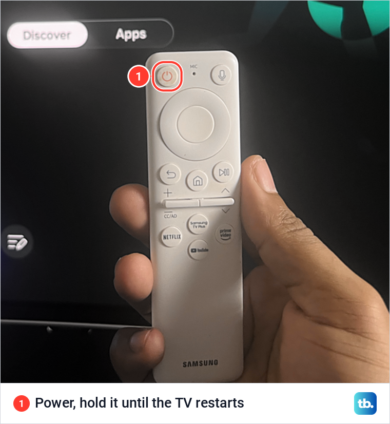 Hand holding a white Samsung remote in front of the Smart Hub screen, with the Power button marked.
