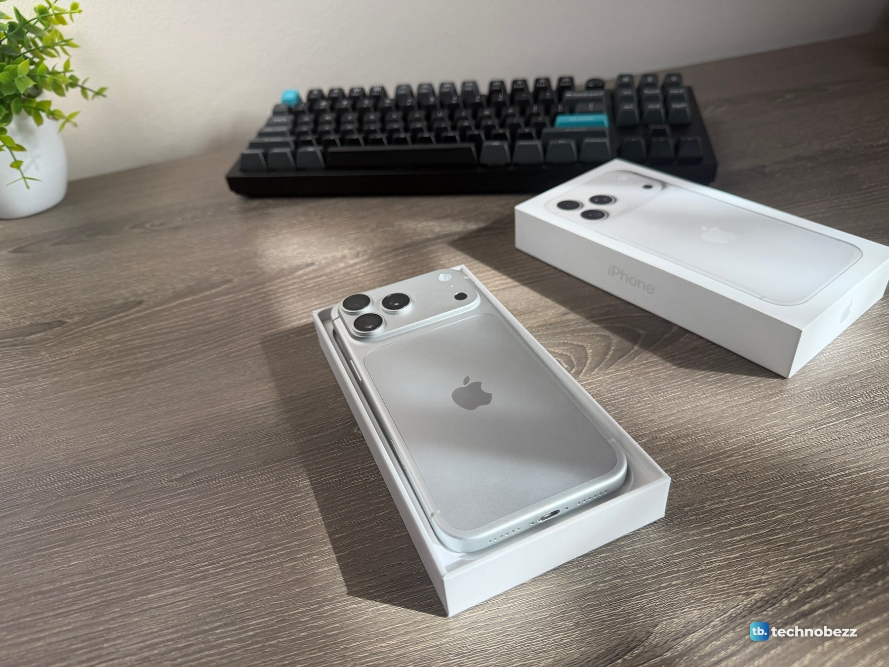 Silver iPhone 18 Pro Max lying face down in its open box tray on a wooden desk, with the box lid and a keyboard behind it