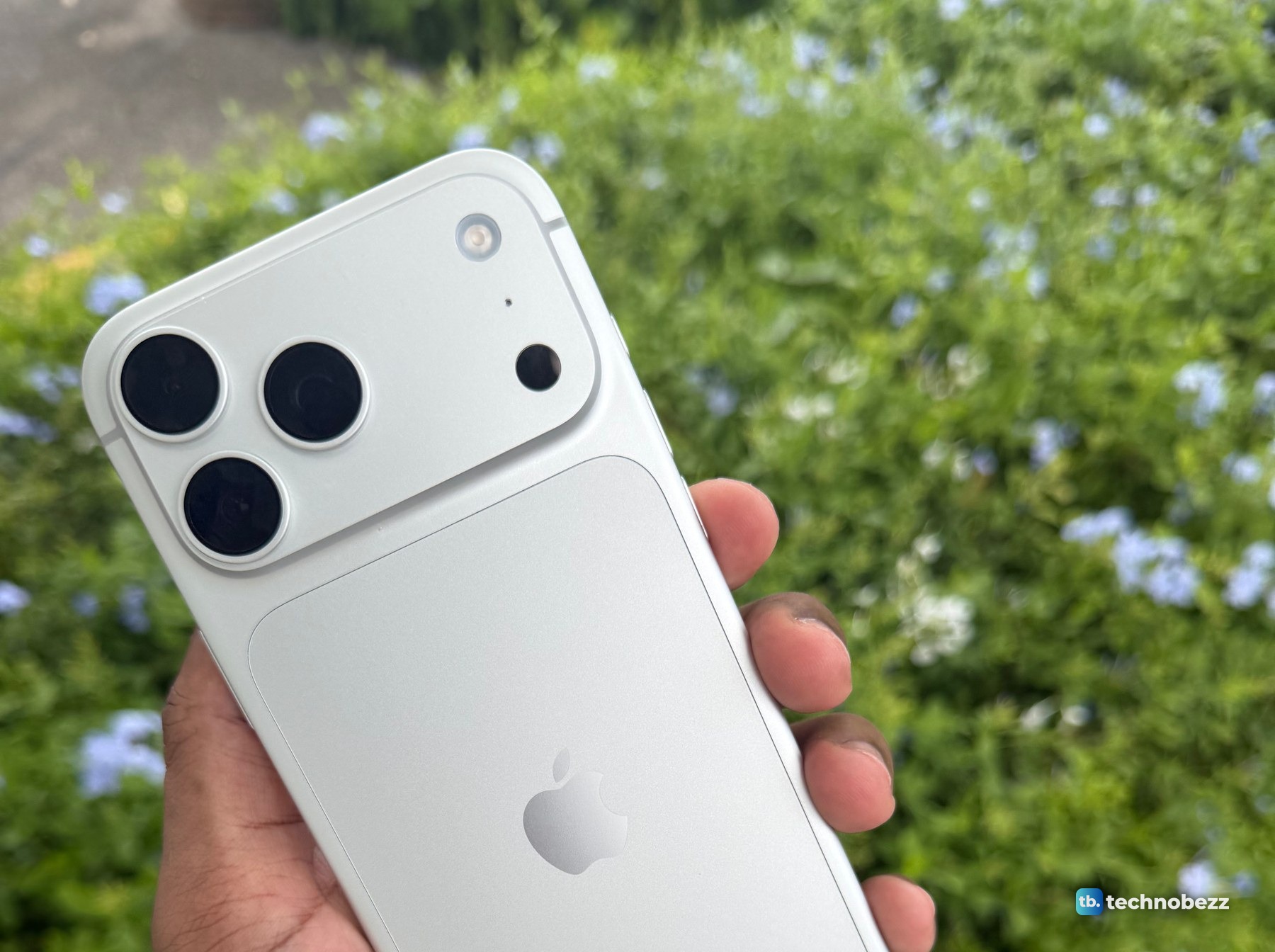 The back of a silver iPhone 18 Pro Max held in a hand, with the camera bar and its three lenses in front of a hedge with blue flowers