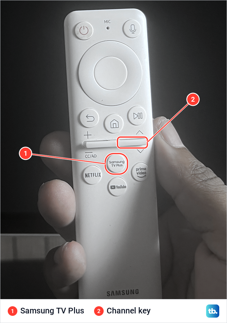 Close-up of a white Samsung remote in a hand with the Samsung TV Plus key and the channel key marked.