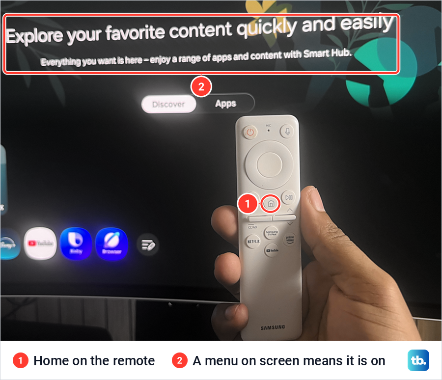 A hand holding a white Samsung remote in front of the Smart Hub menu, with the Home button marked.