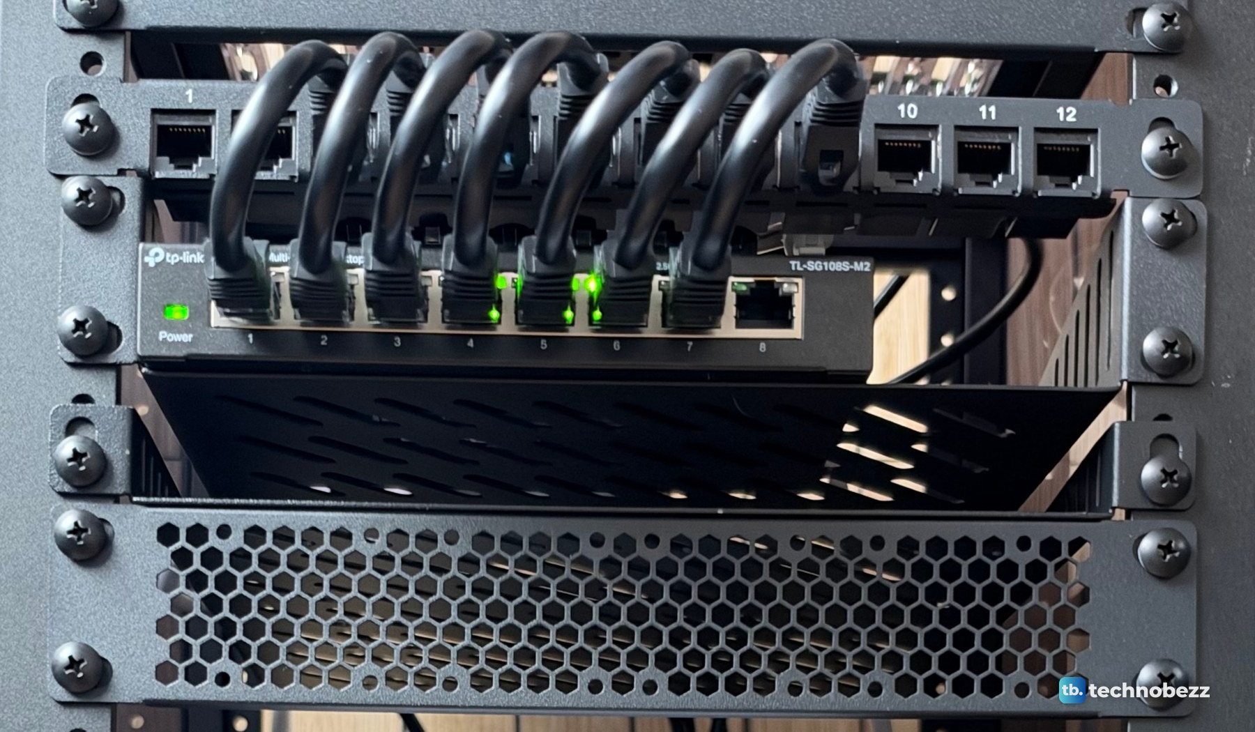 A 12-port patch panel with short black jumper cables looping down into a TP-Link TL-SG108S-M2 switch with green port lights
