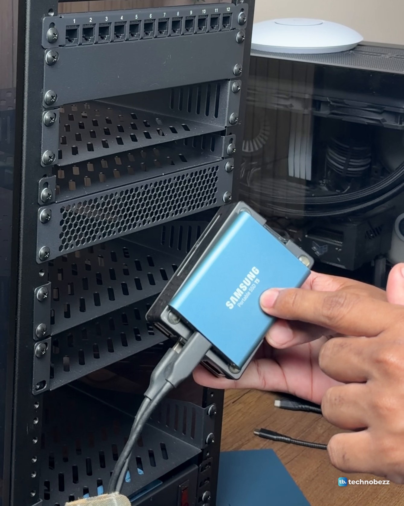 A hand holding a blue Samsung T5 portable SSD on top of a small black Raspberry Pi 5 case, with a cable plugged in