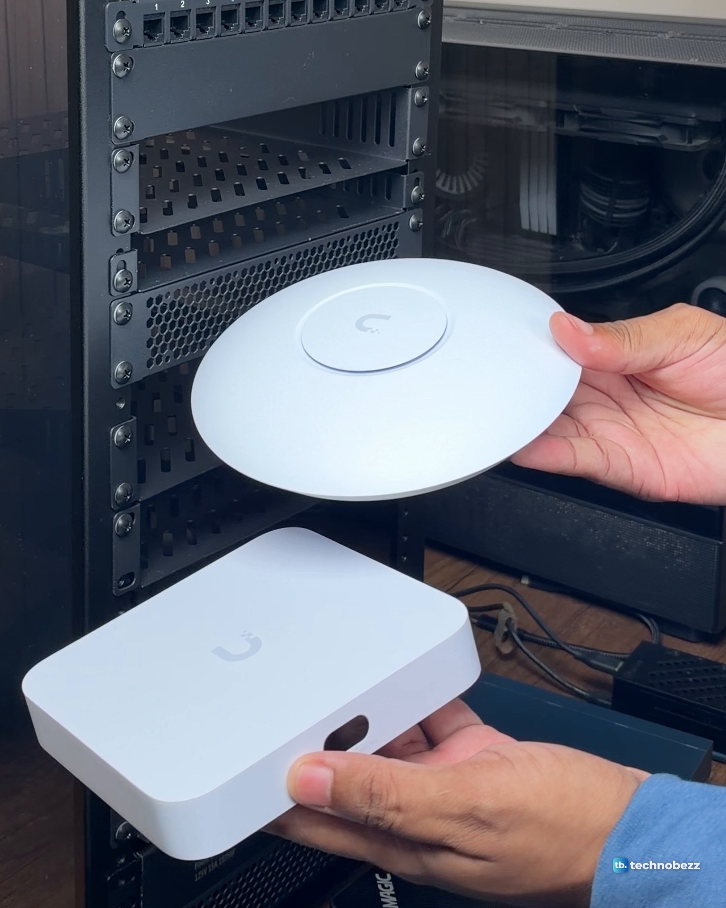 Hands holding a round white UniFi U7 Lite access point above the white UniFi Cloud Gateway Ultra in front of the rack