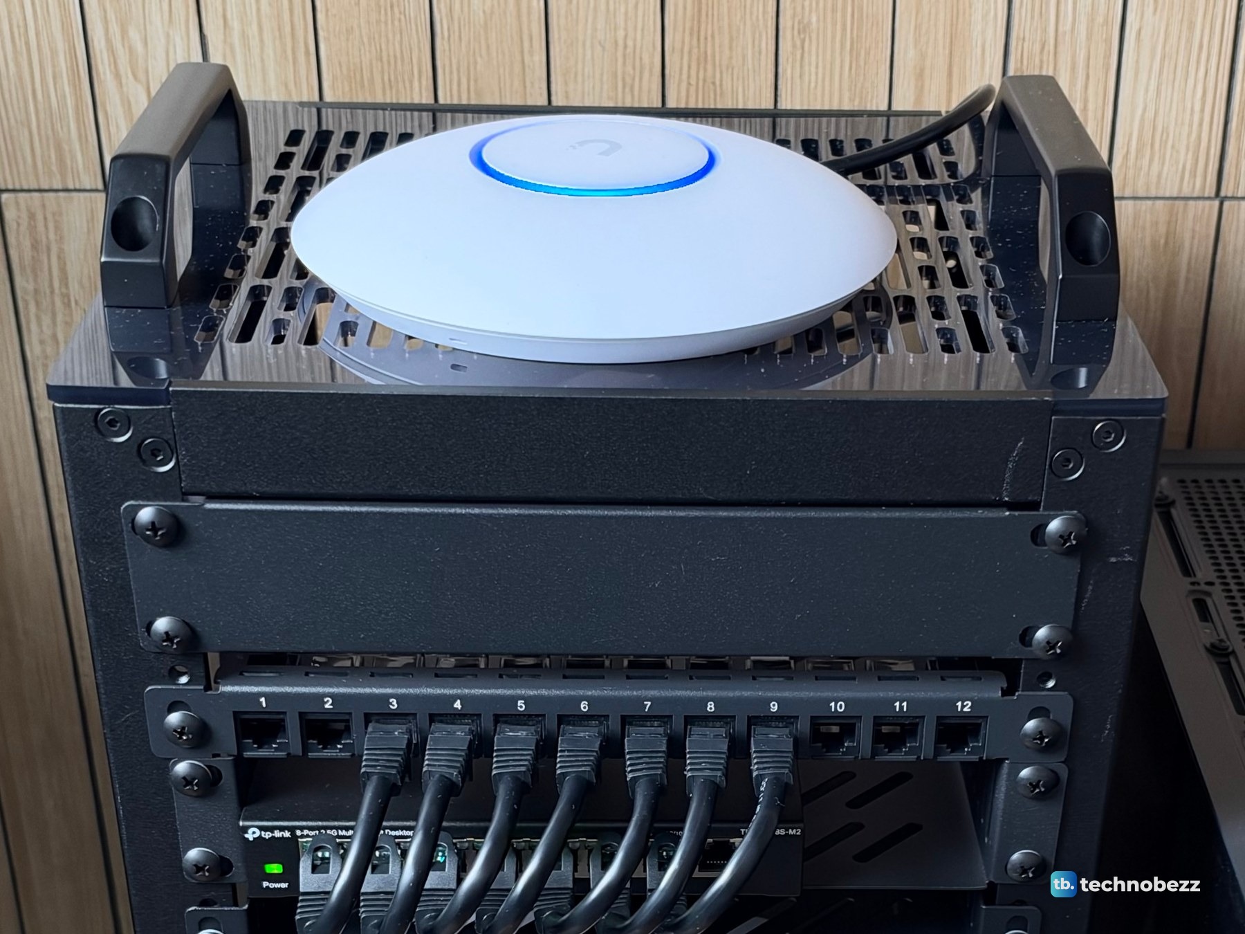 The round white UniFi U7 Lite sitting on the vented top plate of the rack between two black handles