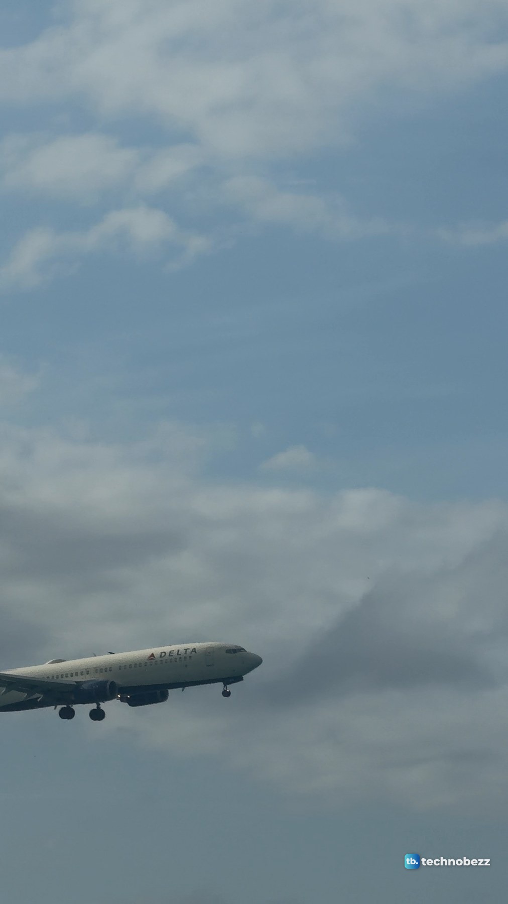 The front half of an airliner with its landing gear down at the left edge of the frame, against cloud and blue sky