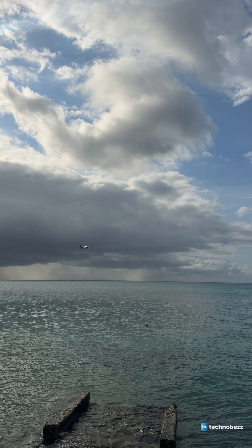 Wide view of the sea under a dark rain cloud with a plane as a small speck in the sky
