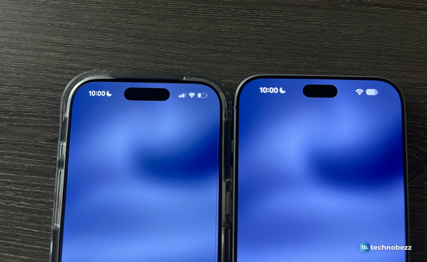 iPhone 17 Pro in a clear case and a silver iPhone 18 Pro Max lying side by side on a dark wood desk, both showing 10:00 and an empty Dynamic Island