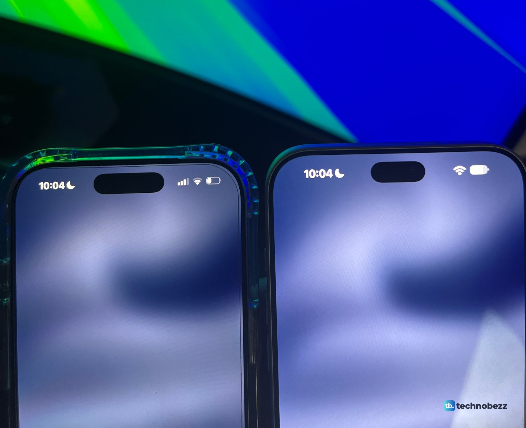 iPhone 17 Pro in a clear case and iPhone 18 Pro Max standing side by side in front of a monitor with a green and blue wallpaper, both showing 10:04 and an empty Dynamic Island