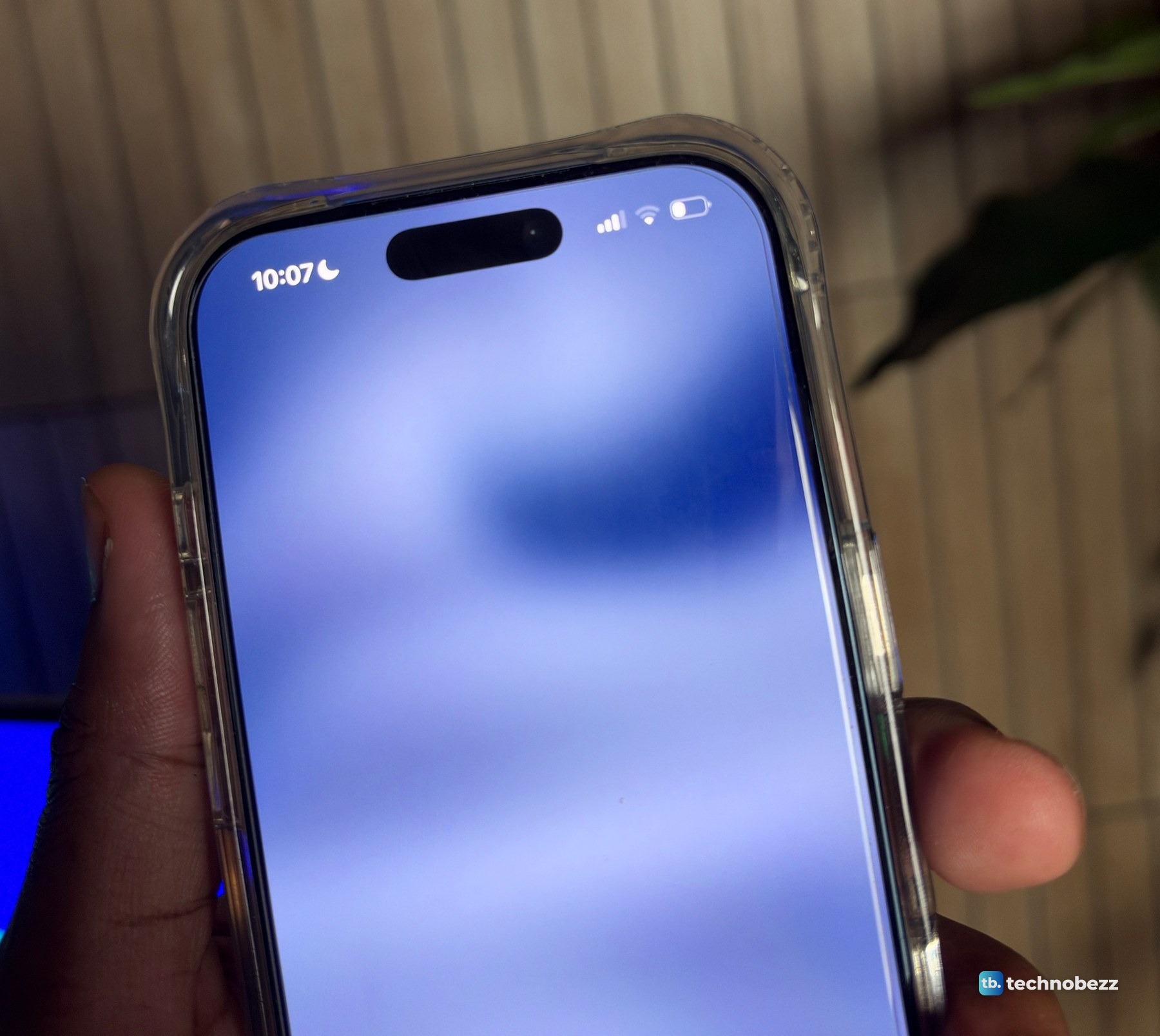 iPhone 17 Pro in a clear case held in one hand in front of a slatted wood wall, the screen showing 10:07 and an empty Dynamic Island