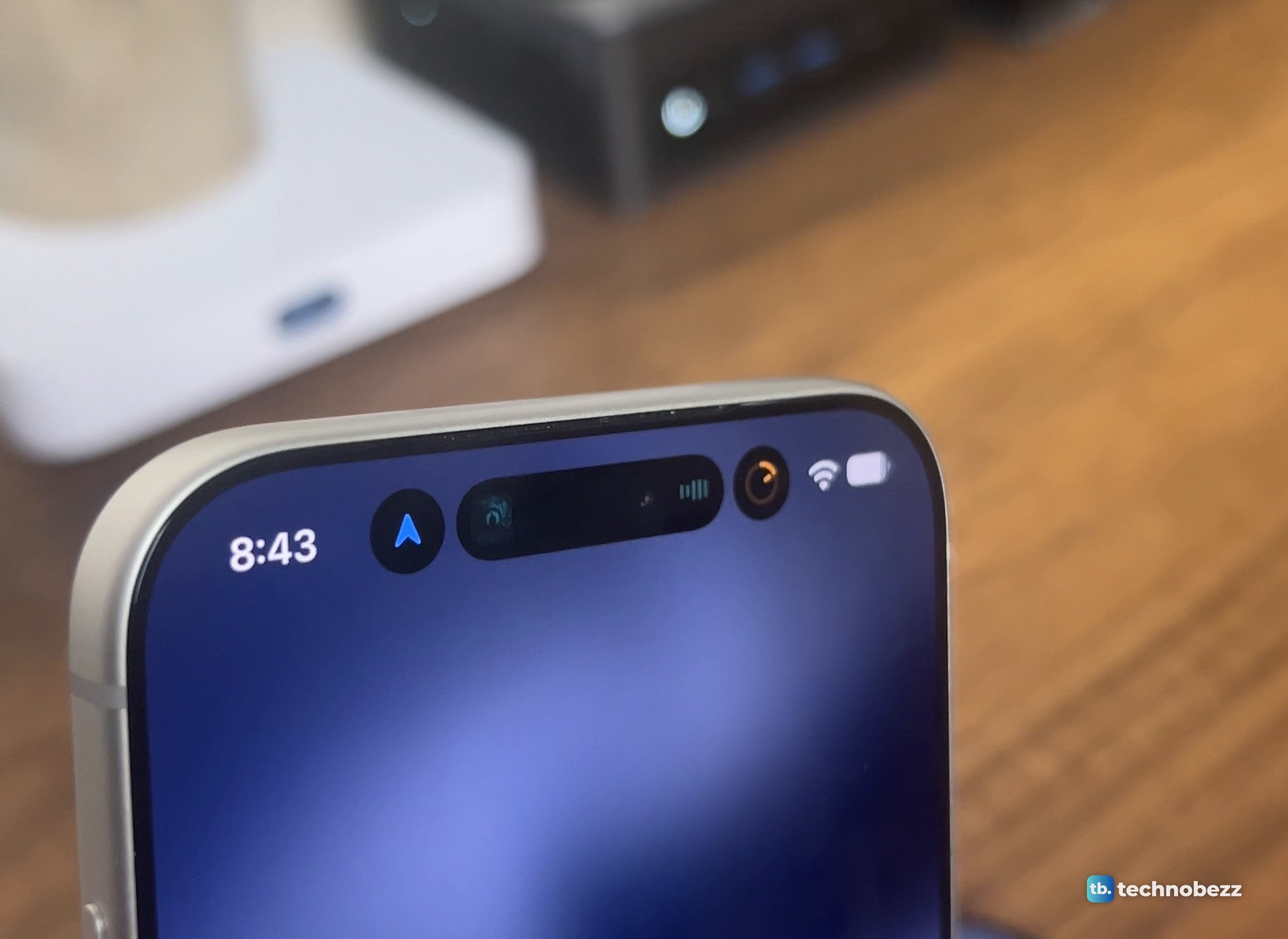 Close view of the top of a silver iPhone 18 Pro Max at 8:43, showing a blue arrow in a round bubble, a dark pill with album art and a waveform, and an orange ring, next to the Wi-Fi and battery icons