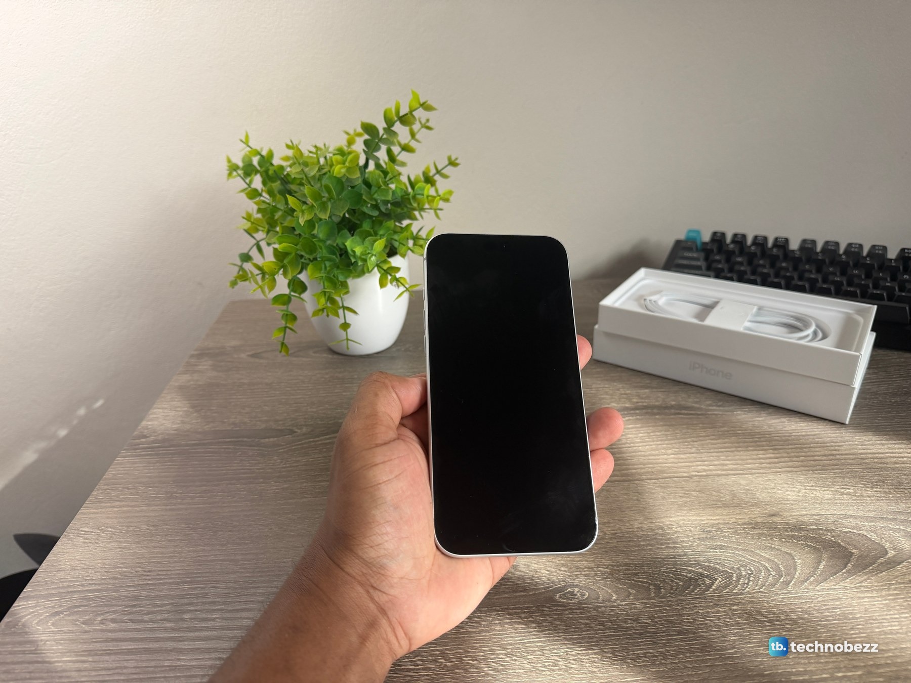 iPhone 18 Pro Max held in one hand with the screen off, and the open box with a white cable on the desk behind it
