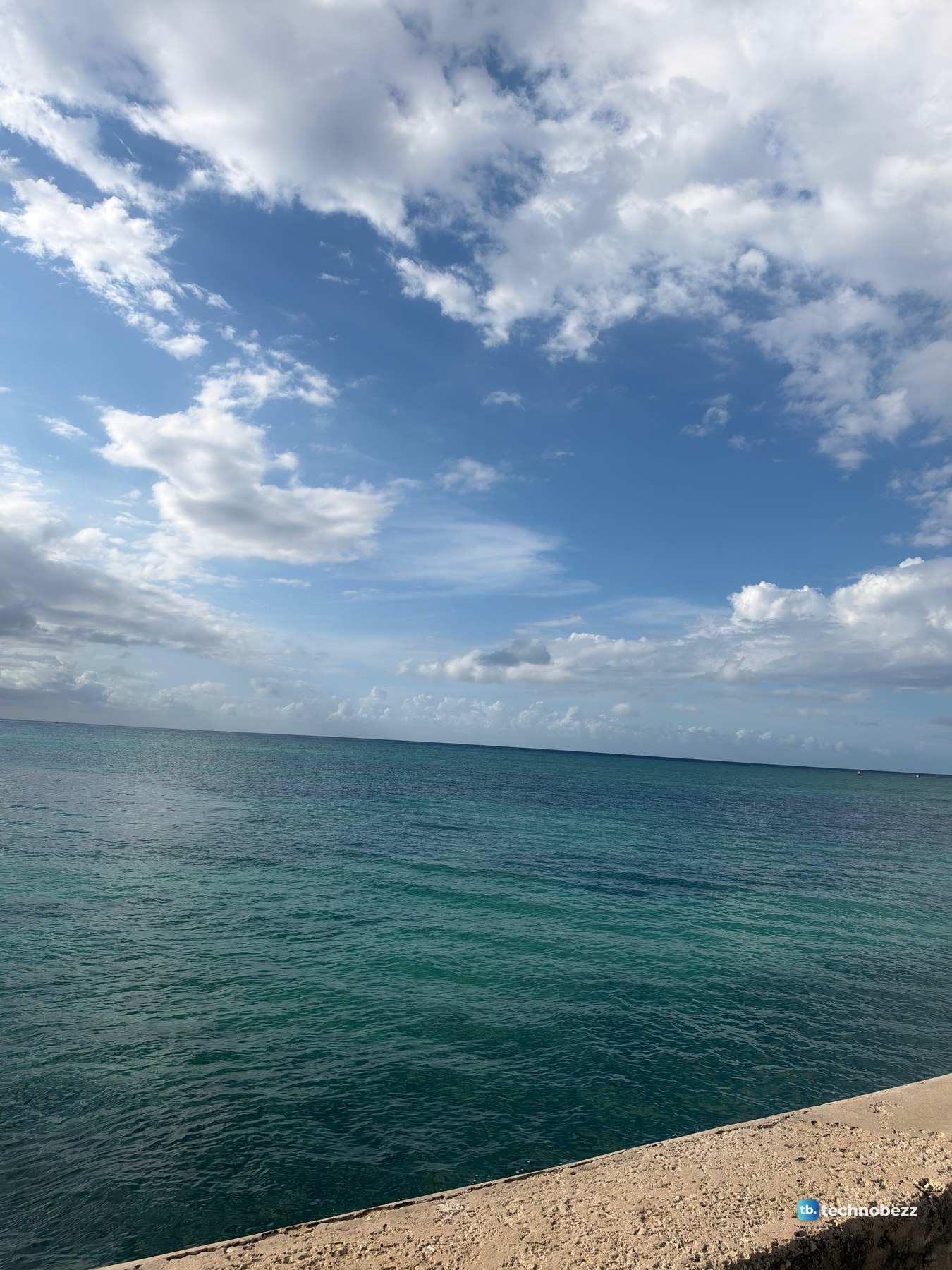 Teal sea under a blue sky with white clouds and the top of a concrete sea wall in the bottom corner, photographed with the iPhone 18 Pro Max