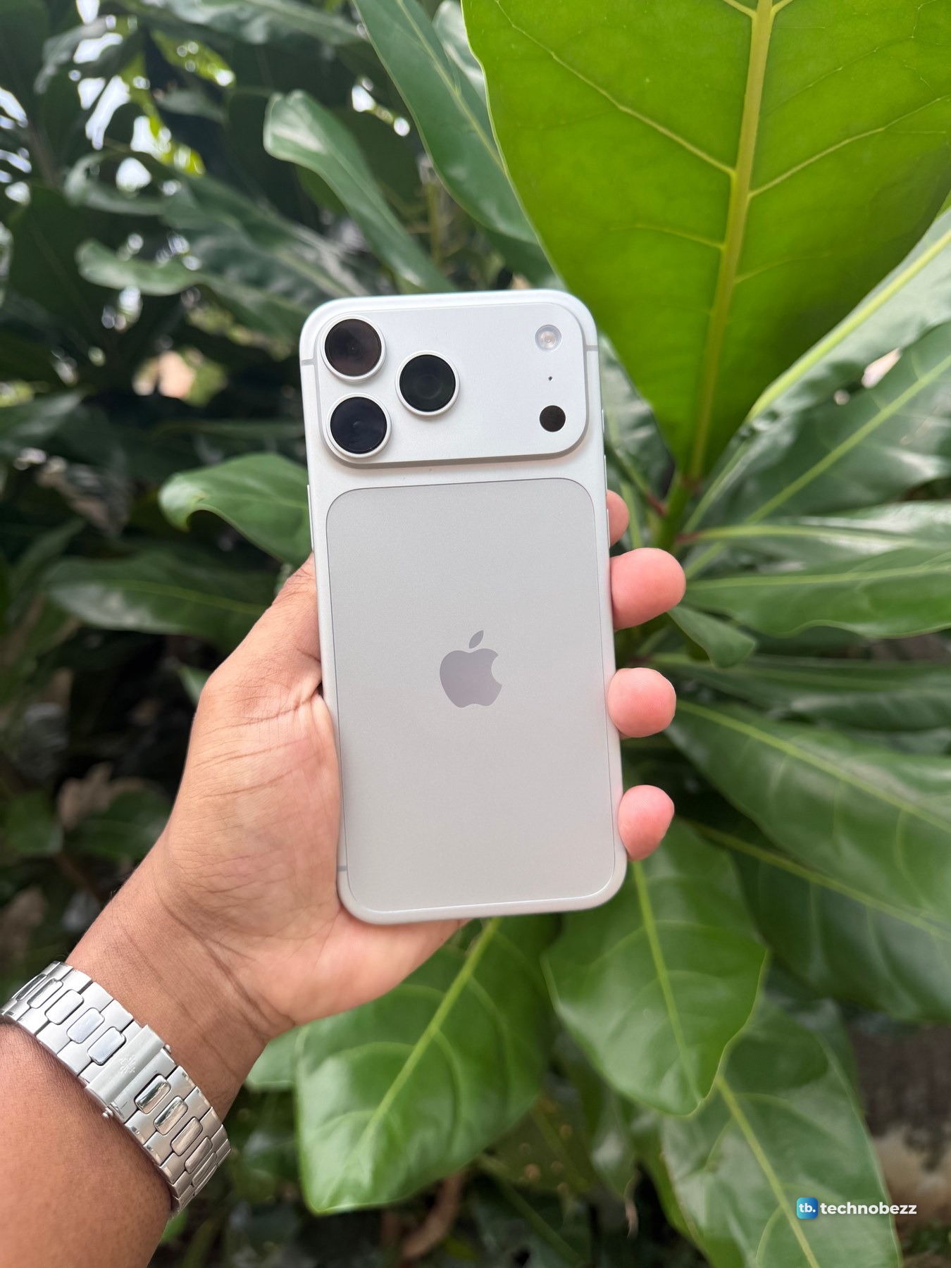 Silver iPhone 18 Pro Max held up in one hand against large green leaves