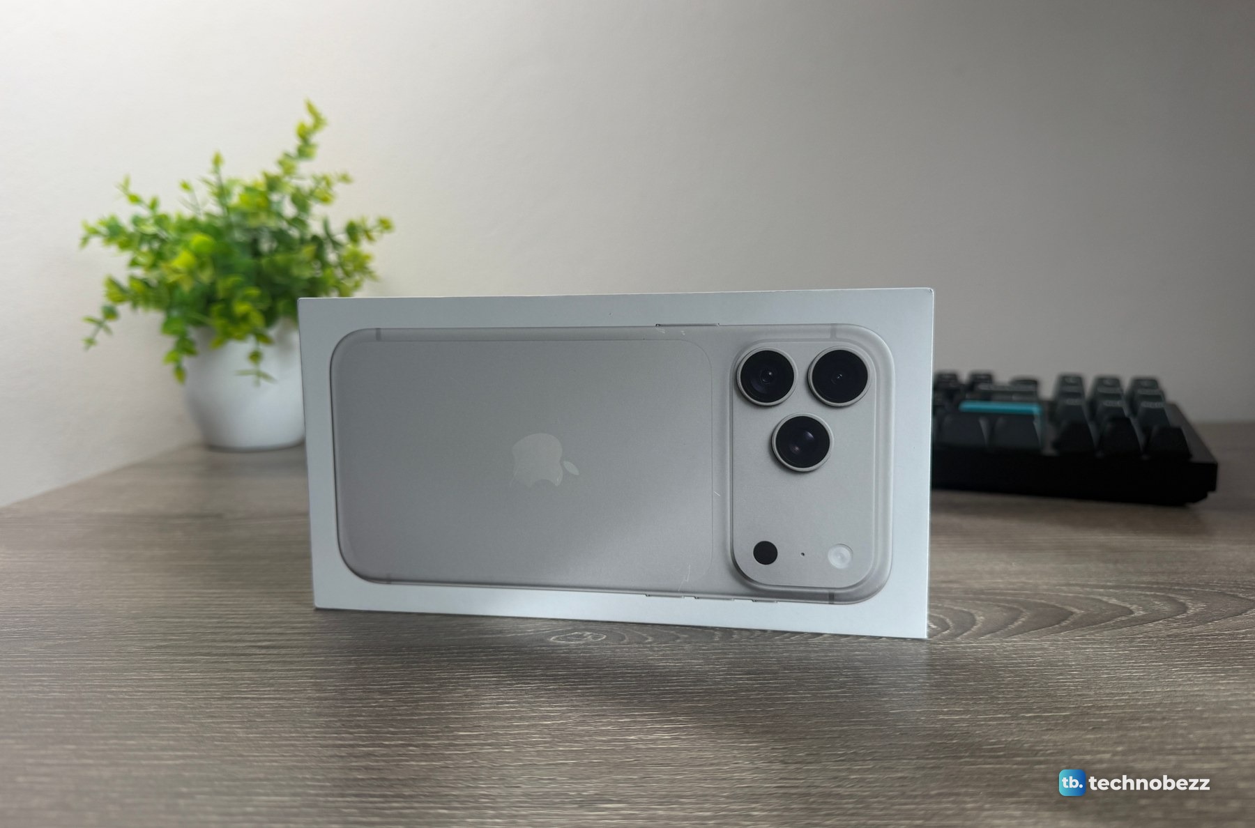 iPhone 18 Pro Max box standing on a wooden desk, with a small plant and a keyboard behind it