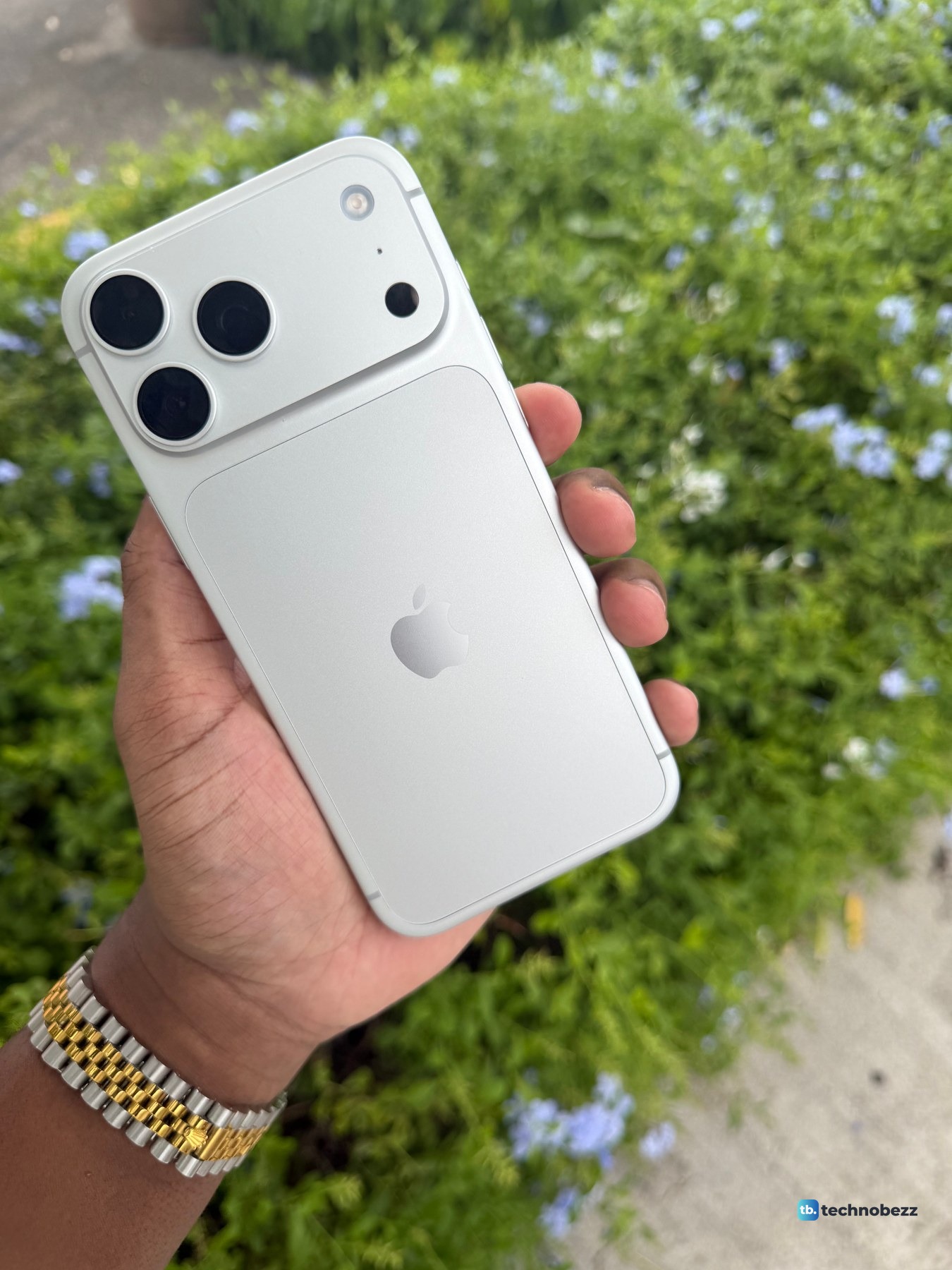 Silver iPhone 18 Pro Max tilted in a hand in front of a green hedge with small blue flowers, the back reading white