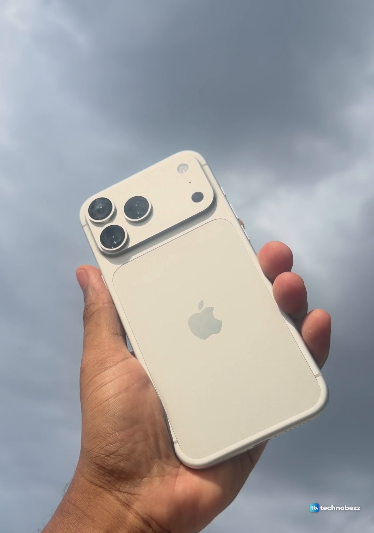 Silver iPhone 18 Pro Max held up against a dark gray storm cloud, the back reading warm cream
