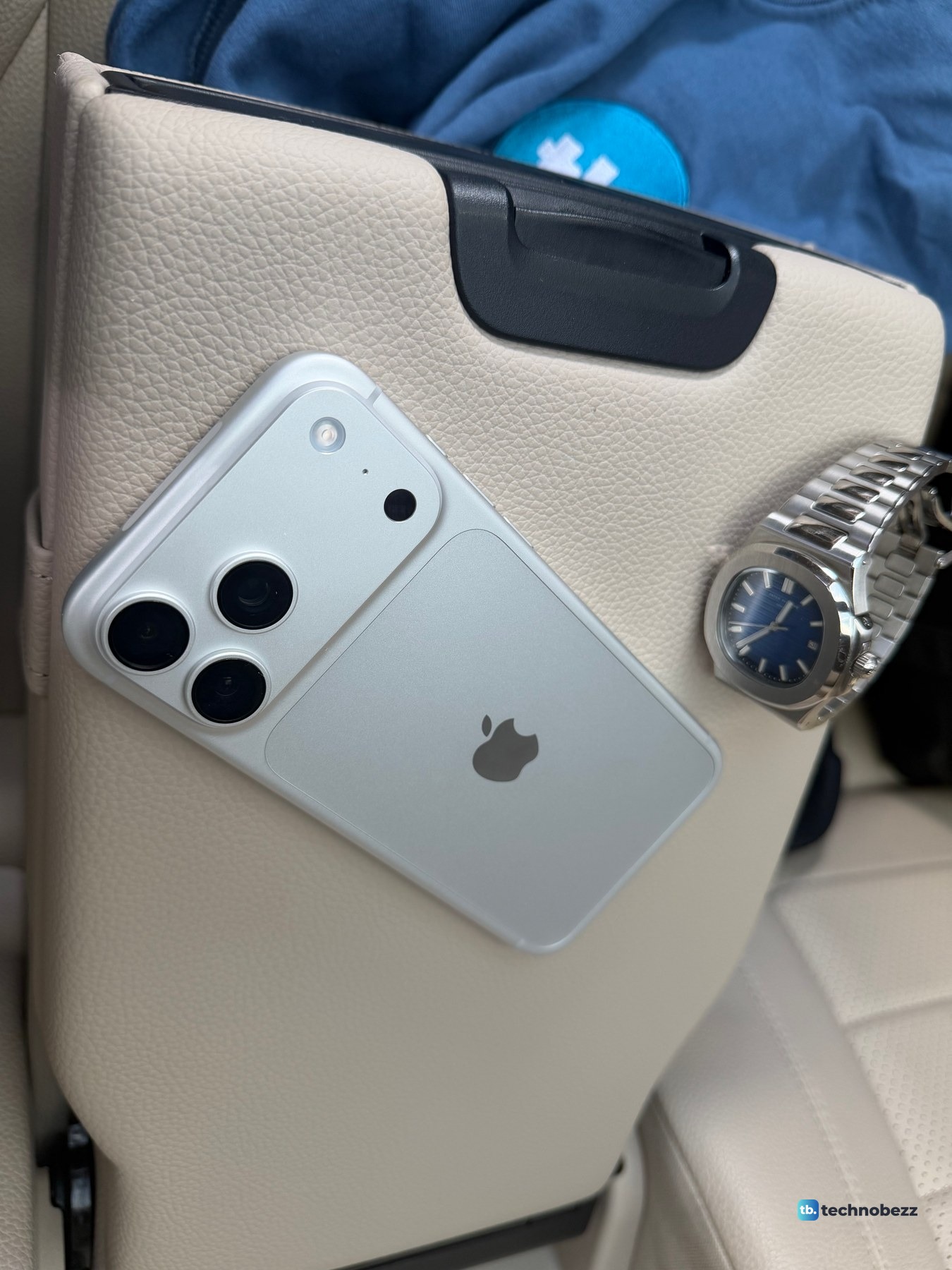 Silver iPhone 18 Pro Max lying on a beige leather armrest next to a steel watch with a blue dial, the back reading an icy silver white