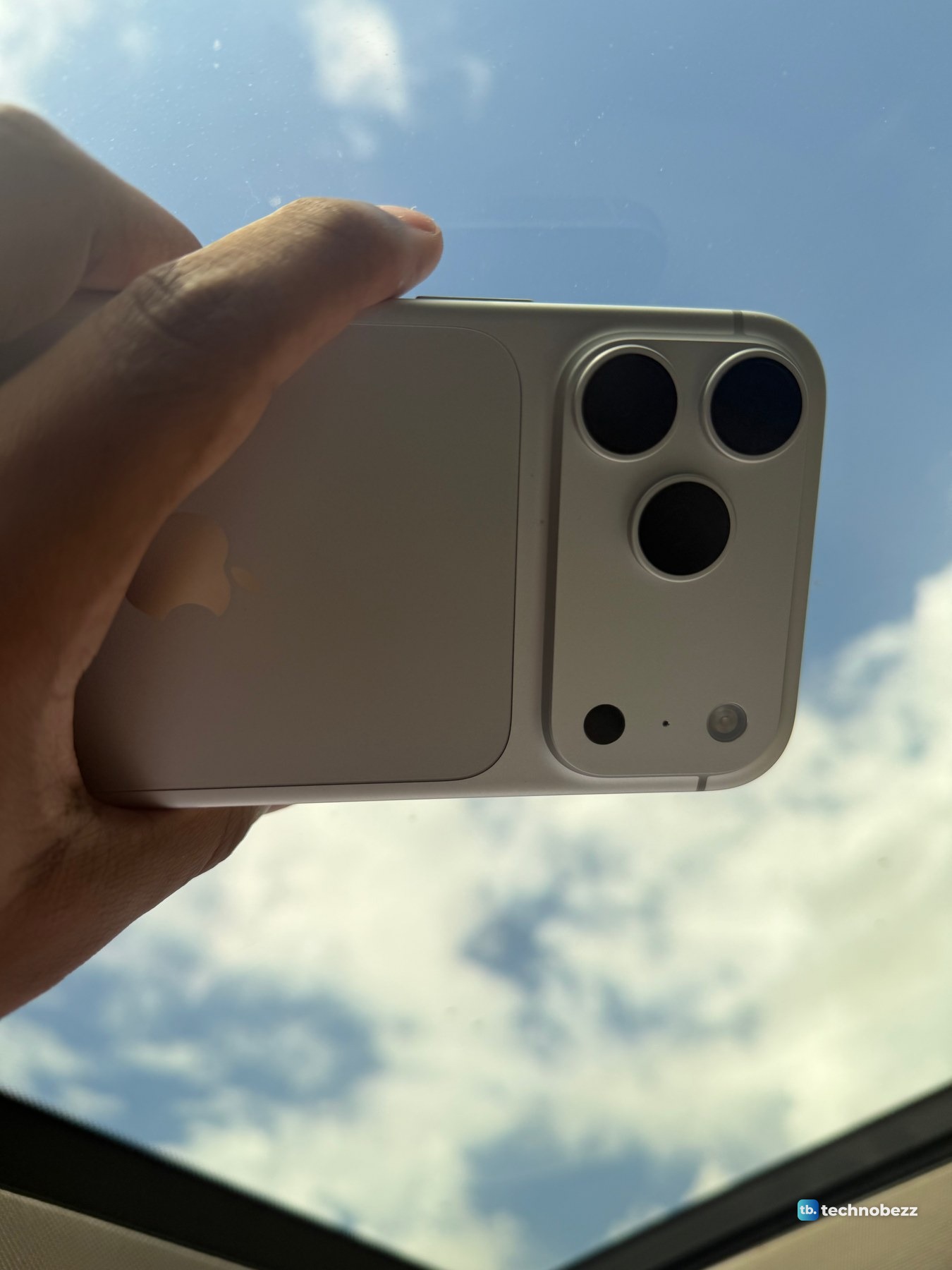 Silver iPhone 18 Pro Max held sideways up to a blue sky with clouds through a car sunroof, the back in shadow and reading warm beige