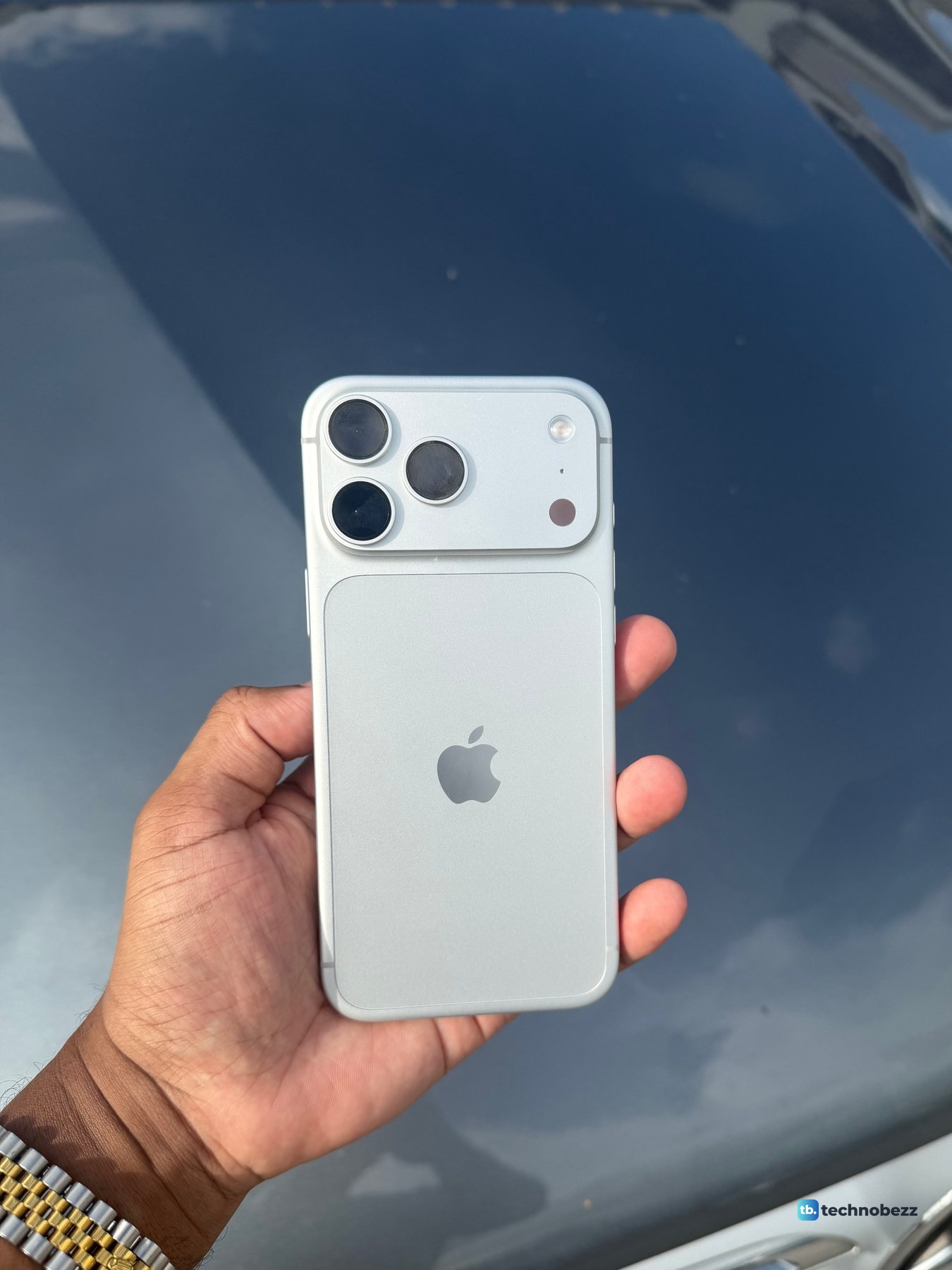 Silver iPhone 18 Pro Max held over a dark gray car hood that reflects the sky and clouds, the back reading almost white