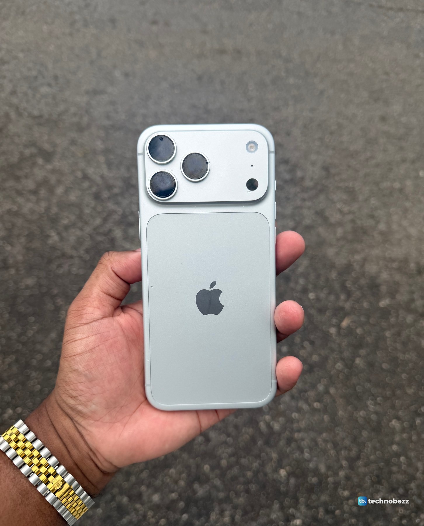 Silver iPhone 18 Pro Max held over plain gray asphalt, the back reading a cool light silver