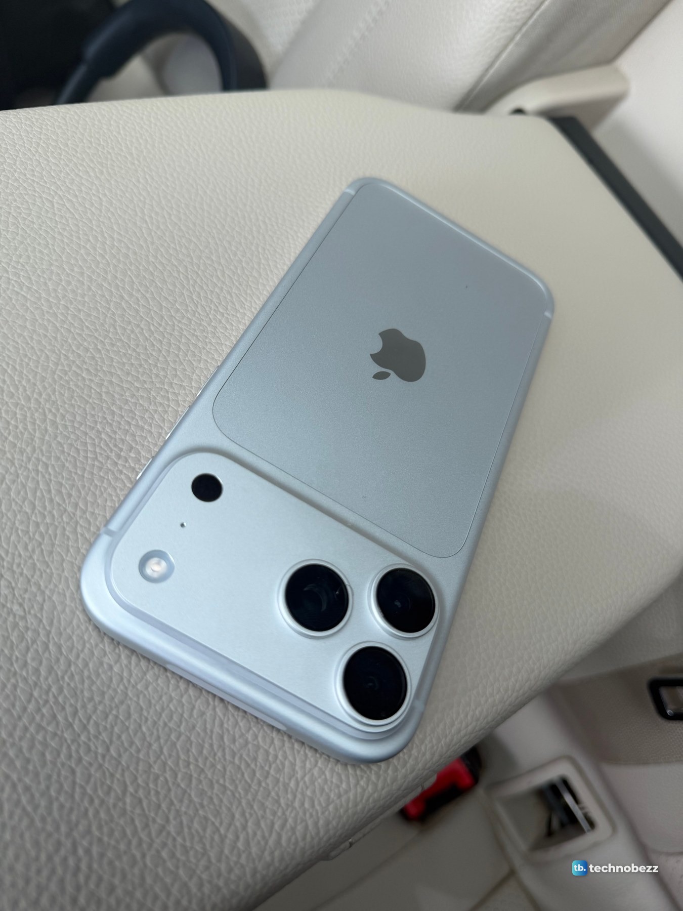 Silver iPhone 18 Pro Max lying on a beige leather armrest inside a car, the back reading pale blue