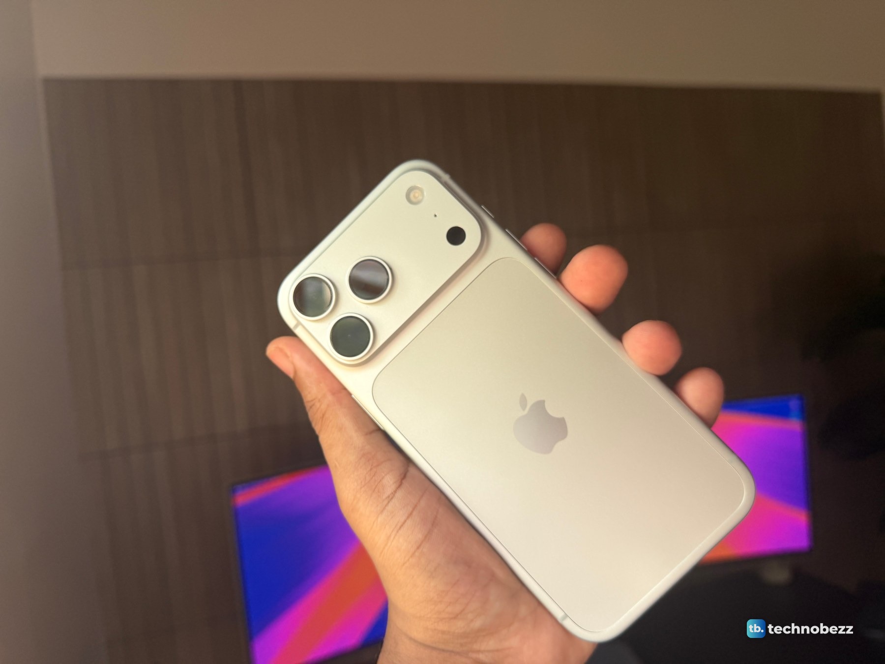 Silver iPhone 18 Pro Max held in front of a dark slat wall and a monitor showing a pink and blue wallpaper, the back reading warm cream