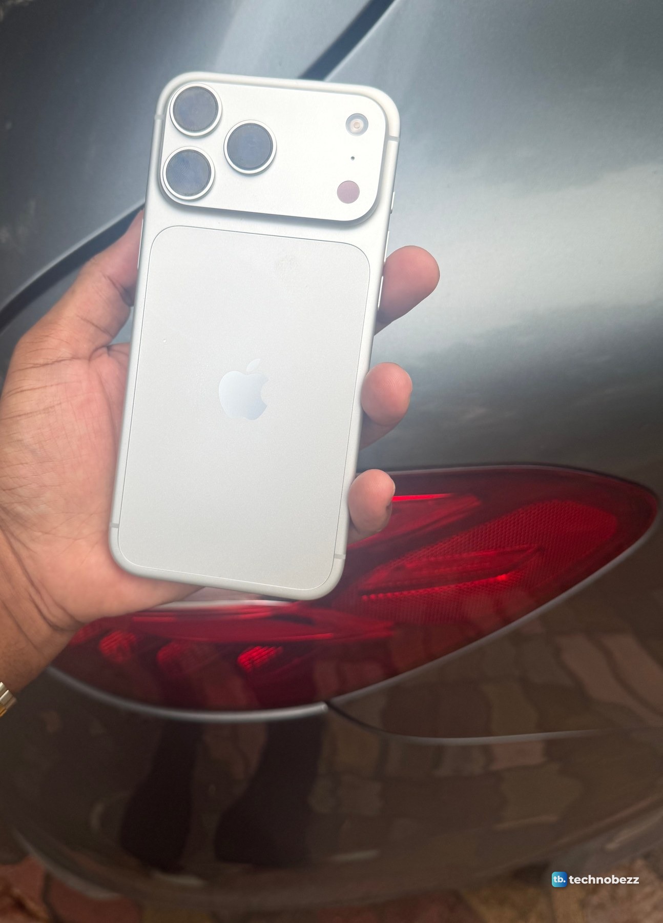 Silver iPhone 18 Pro Max held beside the red tail light of a gray car in soft late light, the back reading warm white