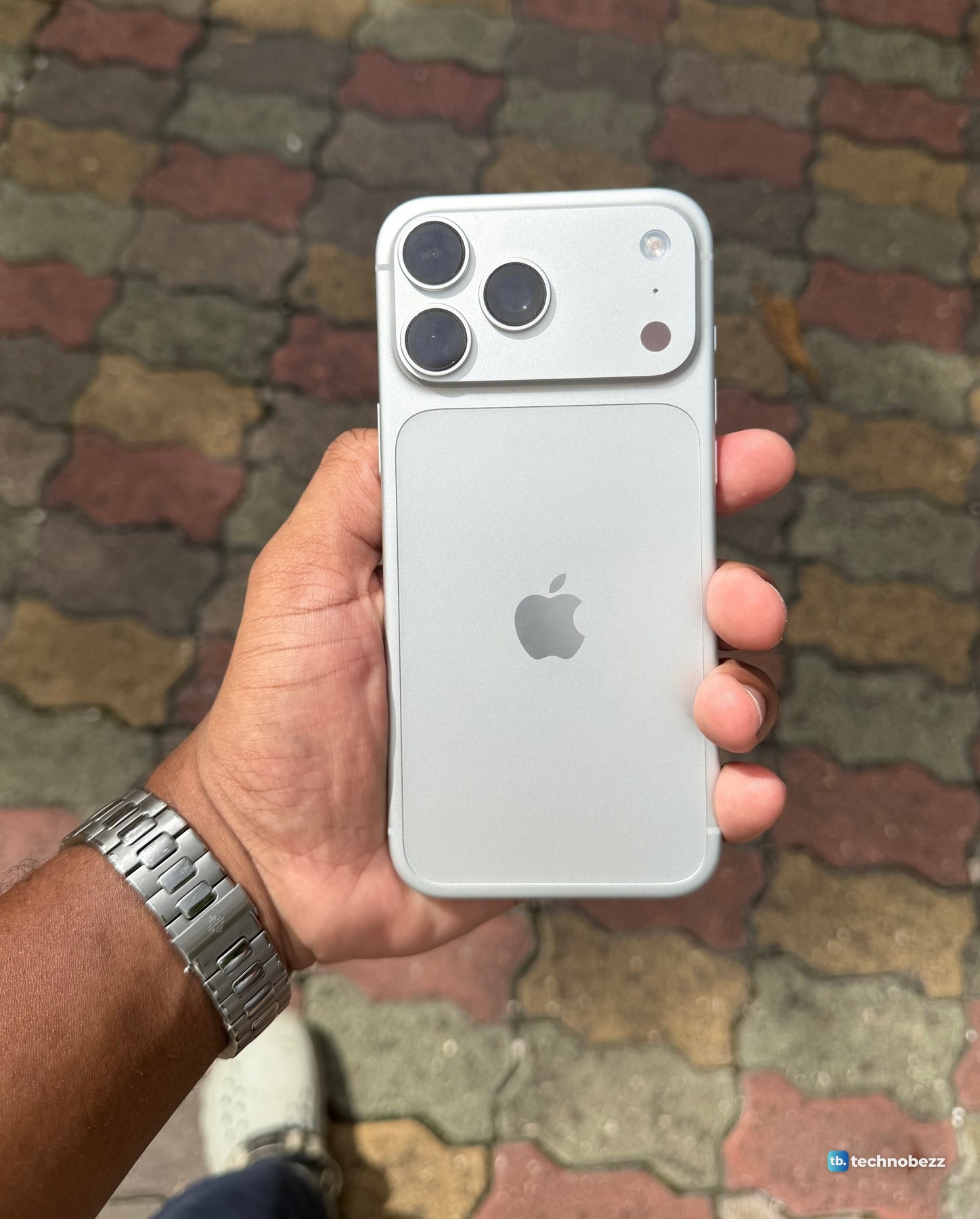 The back of a silver iPhone 18 Pro Max held upright in one hand over colored paving