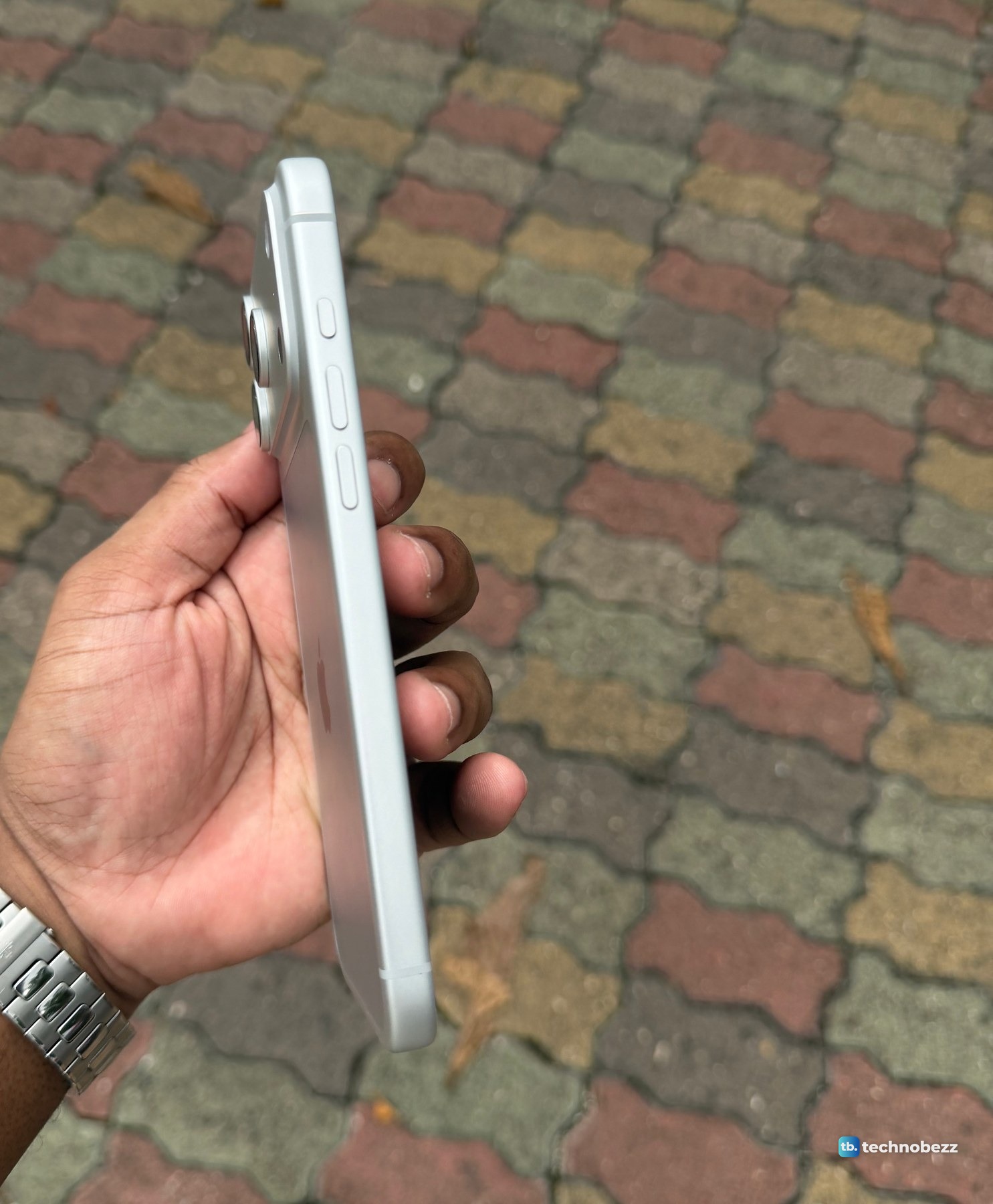 The iPhone 18 Pro Max seen edge-on in an open hand over paving, with three side buttons facing the camera