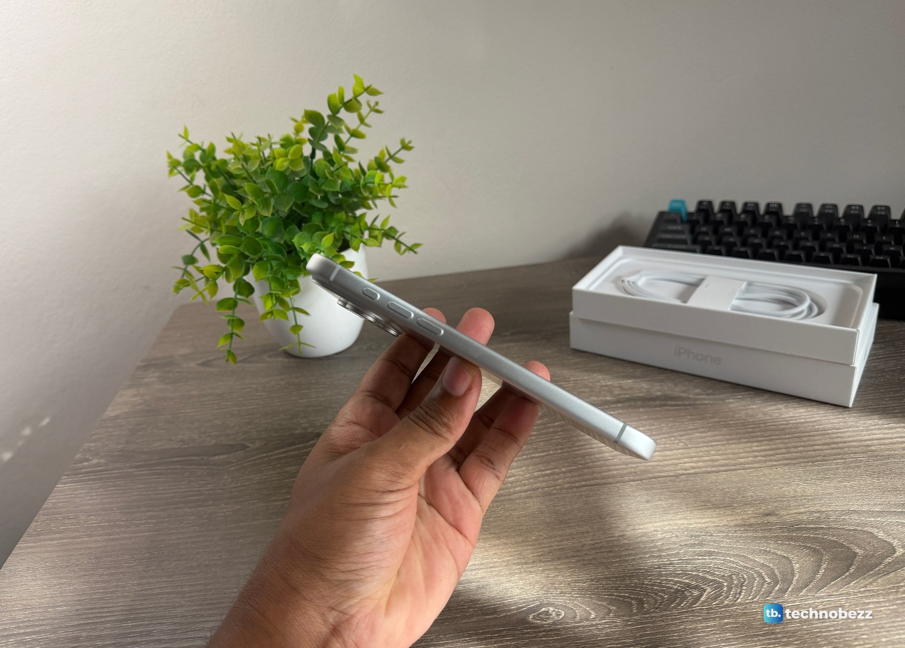 The iPhone 18 Pro Max held flat on its side above a desk, with a plant and the open box with its cable behind