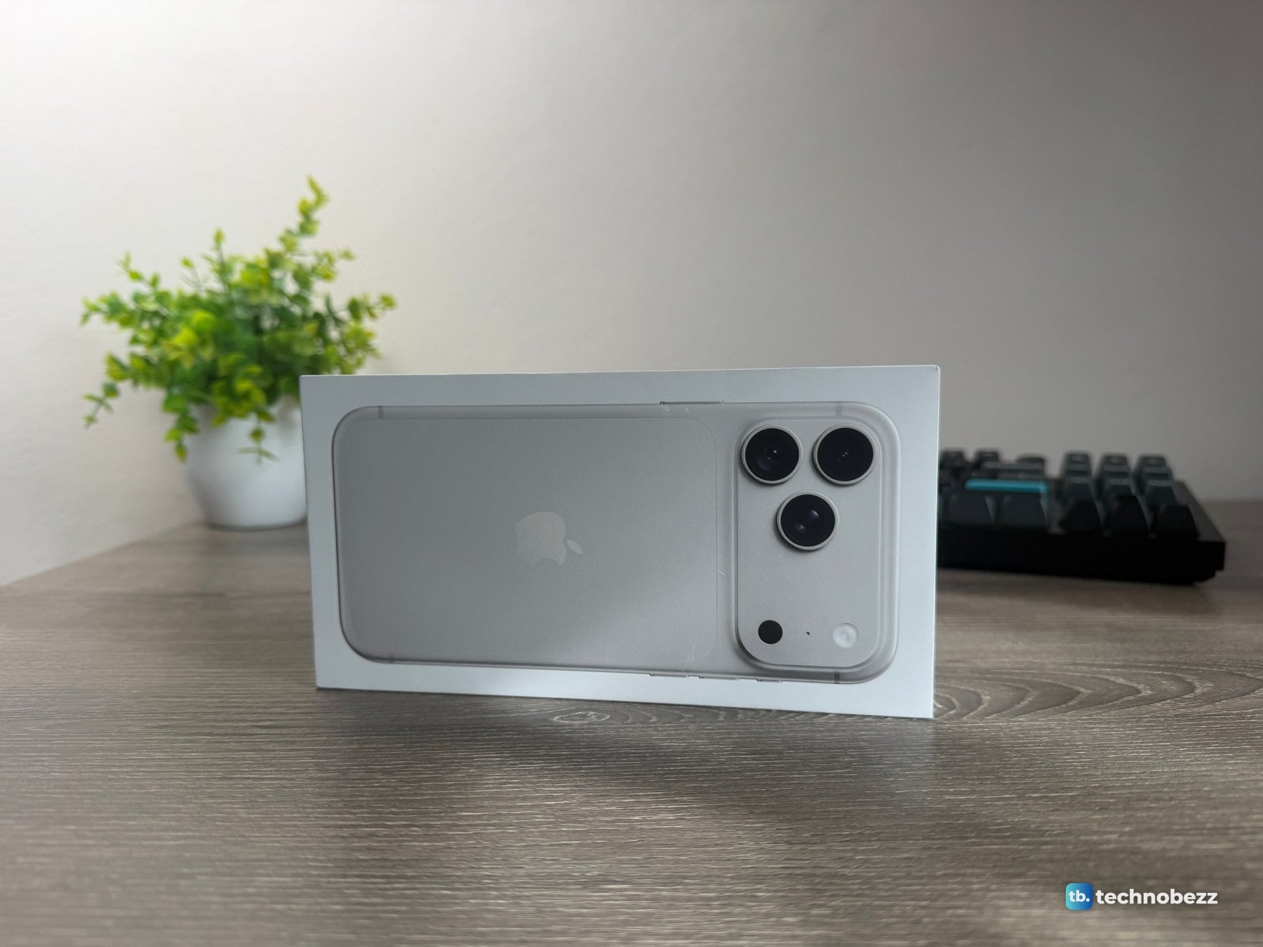 iPhone 18 Pro Max retail box standing on its long side on a desk, the lid showing the silver back of the phone and its three cameras