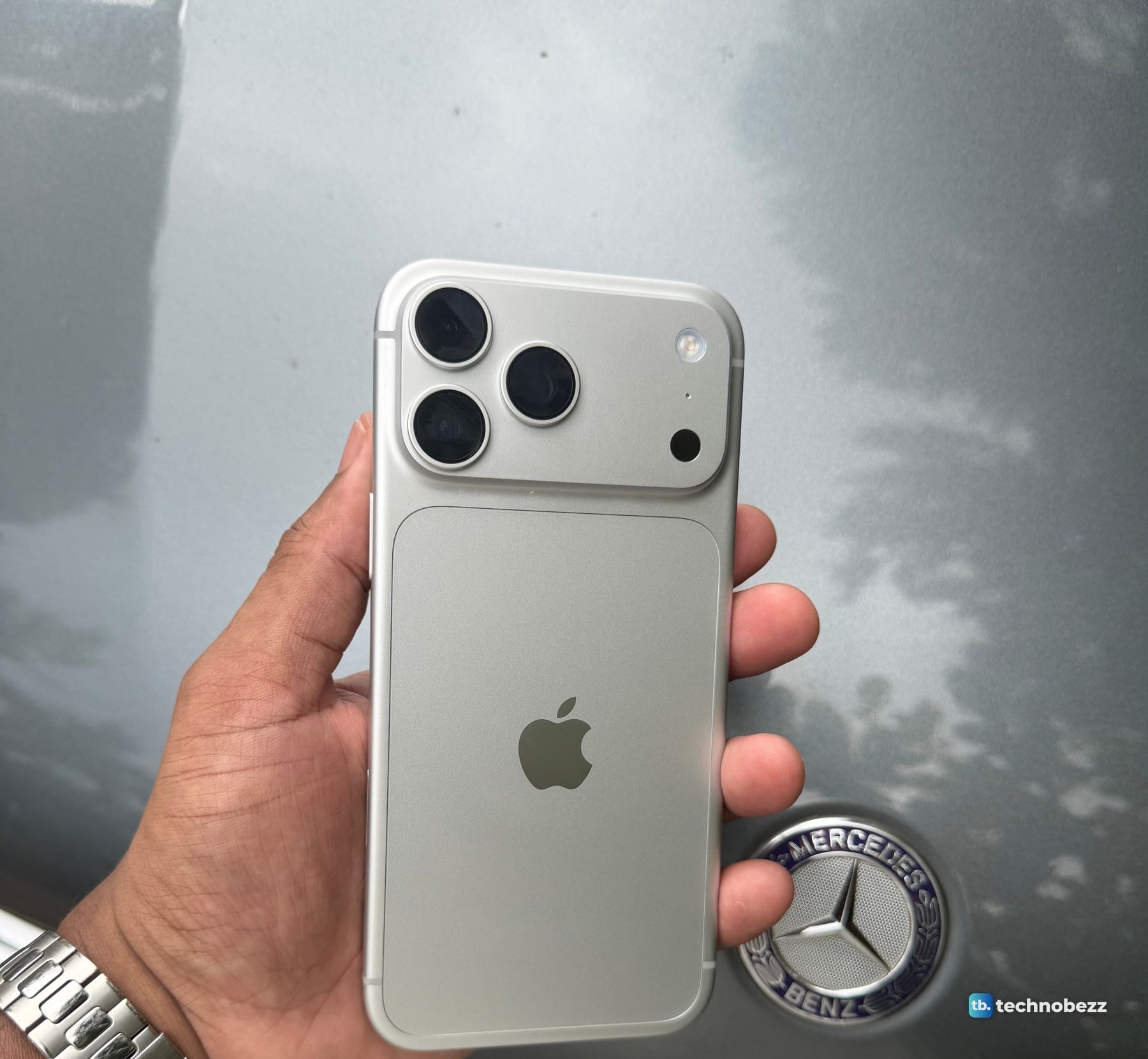 A silver iPhone 18 Pro Max held in a hand beside the Mercedes-Benz badge on a gray car hood